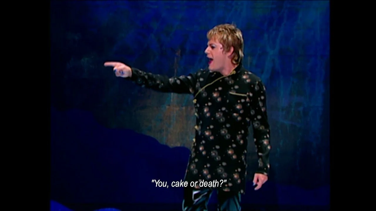 A still from Suzy Eddie Izzard's "Cake or Death" joke. Suzy, a white person with short blond hair is pointing at an imaginary person. The subtitle reads, "You, cake or death?"
