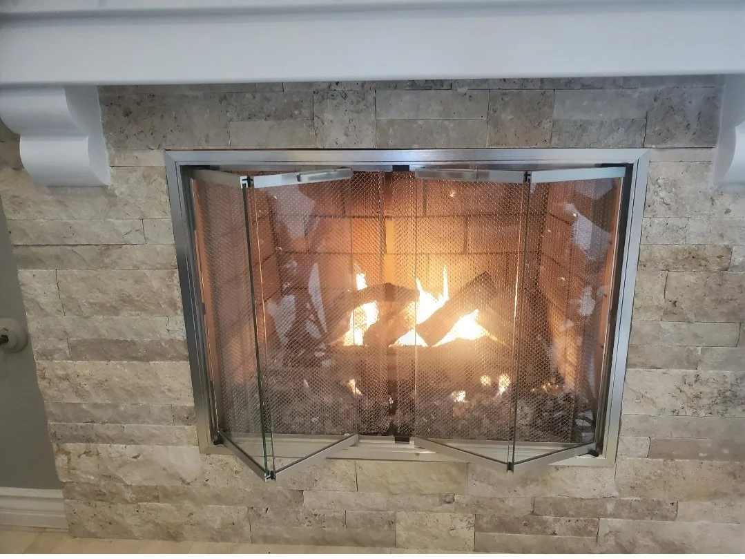 A fireplace with a fire burning inside, enclosed by a glass and metal screen, built into a wall made of beige stone bricks.