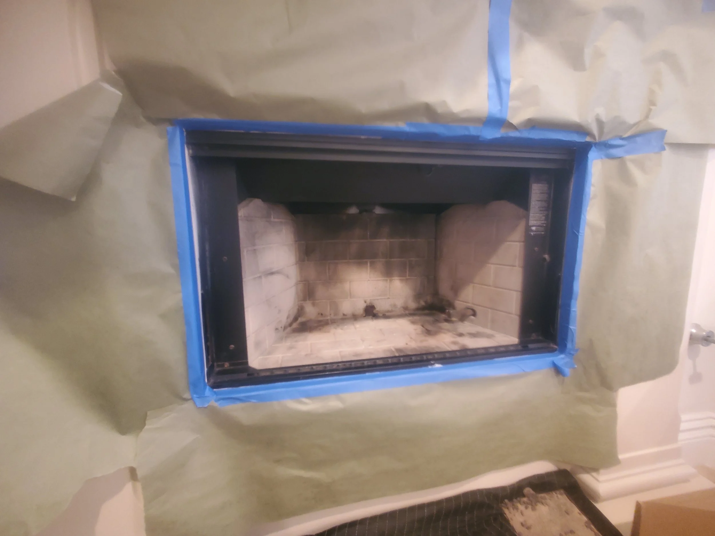 Unfinished fireplace surrounded by painter's paper and tape, with soot and ash inside the firebox.
