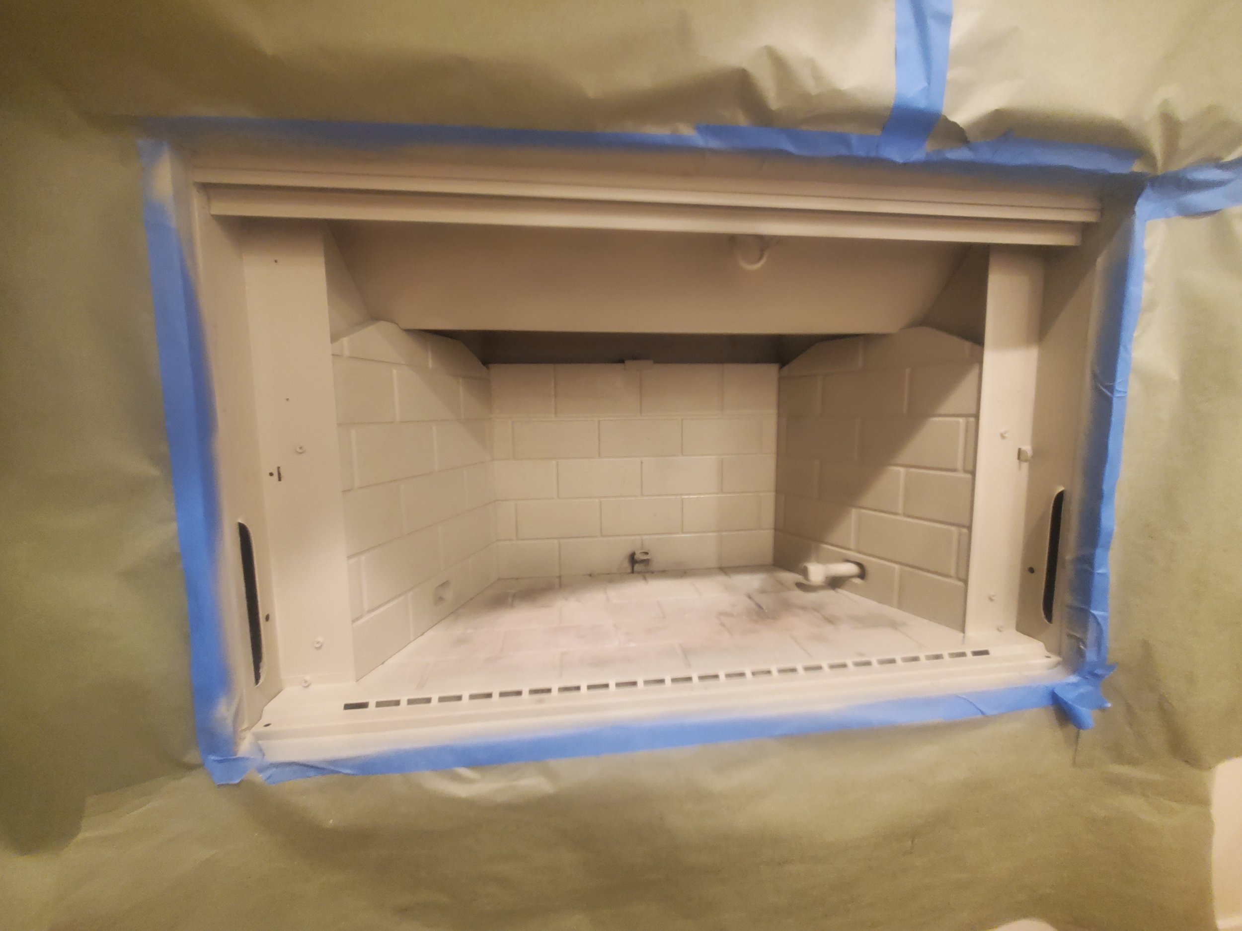 Unfinished fireplace with white brick interior and white mantel, surrounded by painter's tape and paper for painting or renovation.