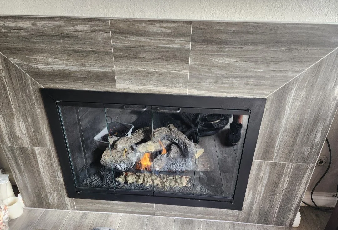 A fireplace with a glass door, showing artificial logs and a small flame inside, surrounded by beige and brown tiled wall.