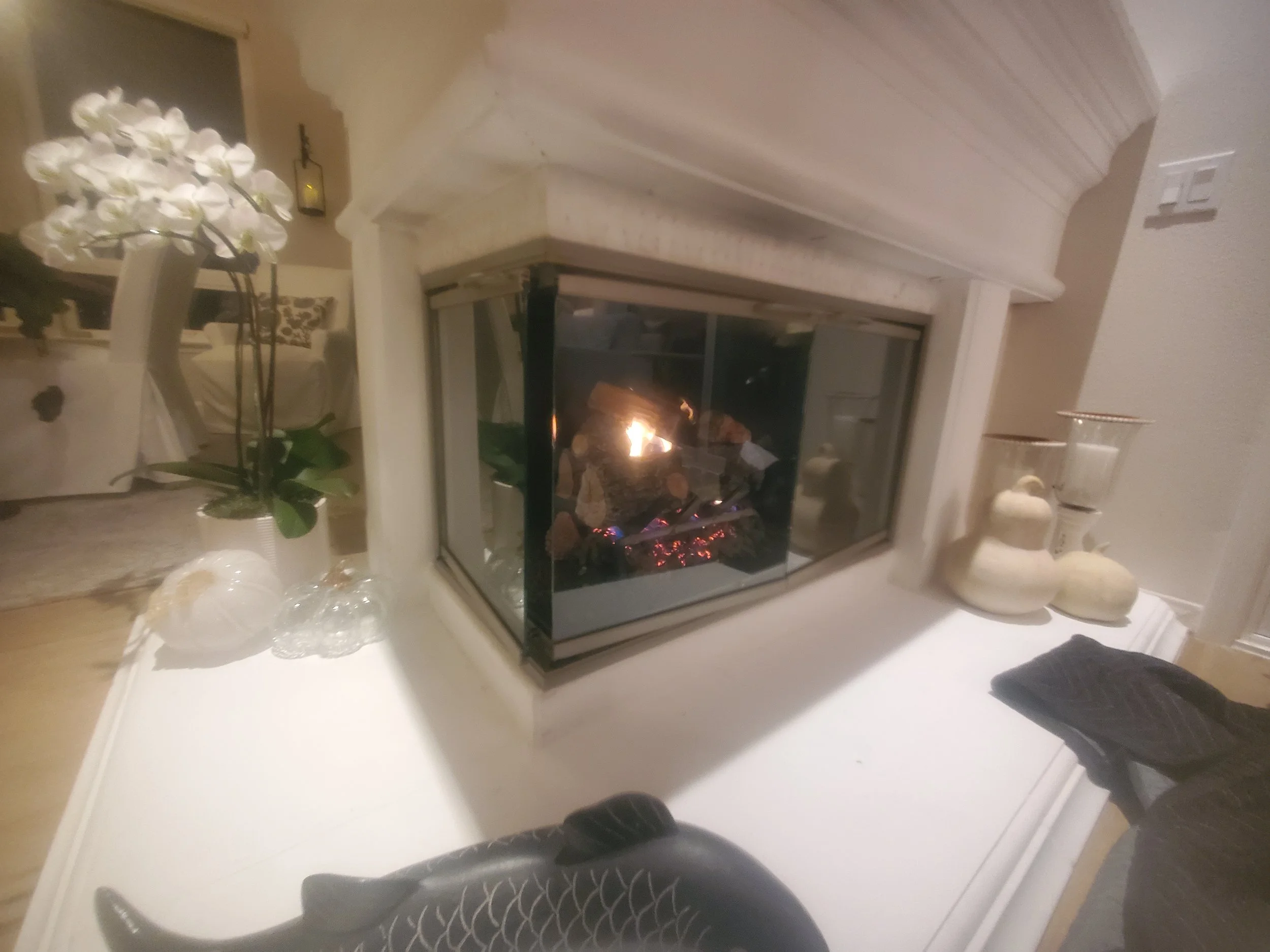 Indoor scene featuring a white fireplace with a lit fire, a white armchair in the background, a side table with glass decor, and a white orchid plant on the left. To the right, there are decorative stones and vases on a surface.
