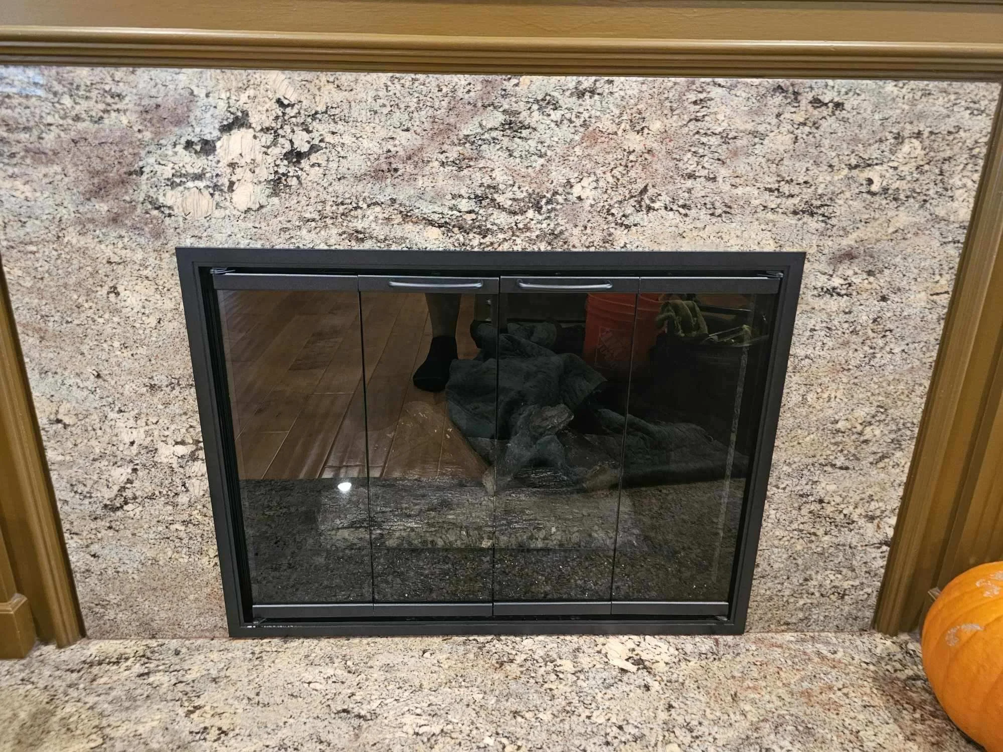 A fireplace framed with black metal surrounded by a beige, speckled granite wall and base. Reflections show a person wearing black socks, a grey shirt, and jeans, with a pumpkin on a side table nearby.