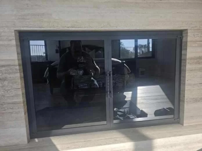 Reflection of a person taking a photo in a sliding glass door, with a car and window visible inside a room behind.