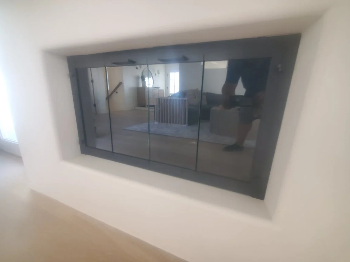A modern built-in fireplace with a glass front, set into a white wall, reflecting a living room with furniture and a person taking the photo.