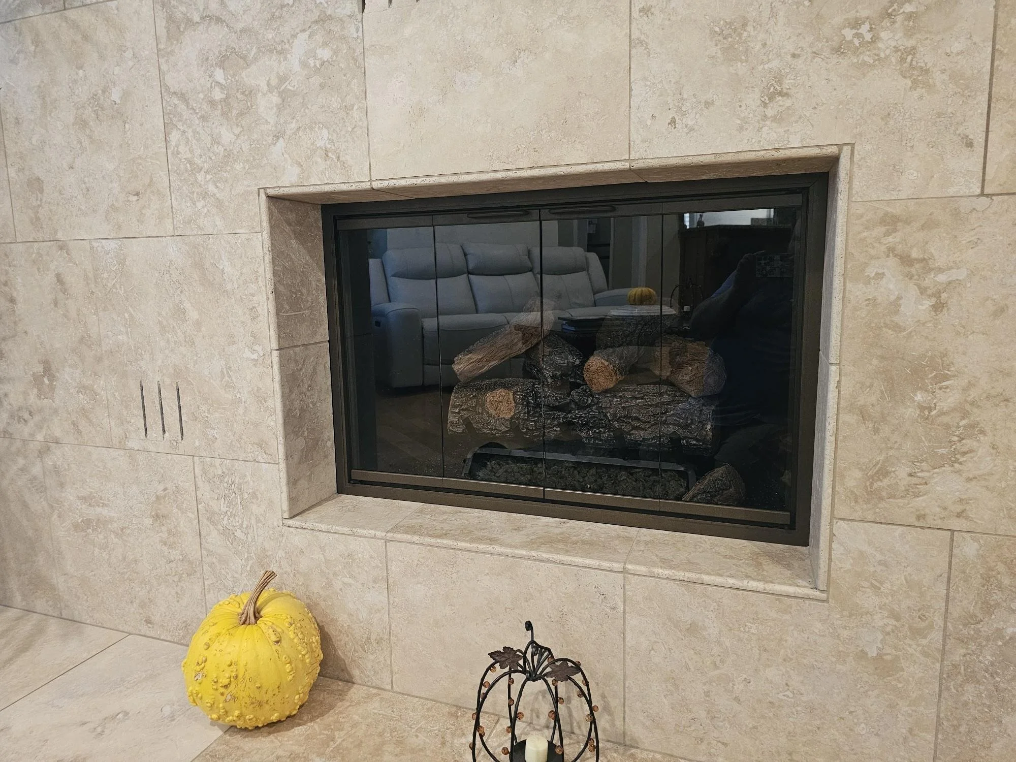 A modern fireplace with glass doors built into a beige stone wall. On the floor in front of it, there is a yellow pumpkin and a small decorative metal pumpkin-shaped stand with a candle inside.