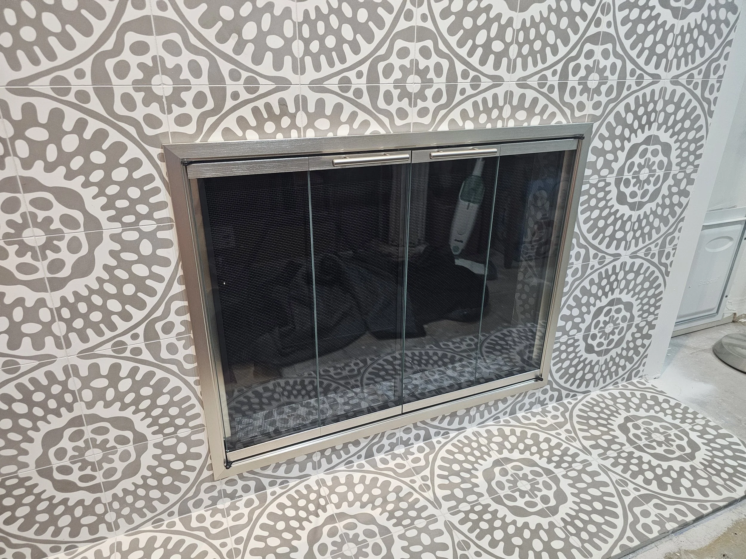 A modern fireplace with a glass door and metal frame installed in a wall covered with patterned gray and white tiles, with matching tiles on the floor in front of it.