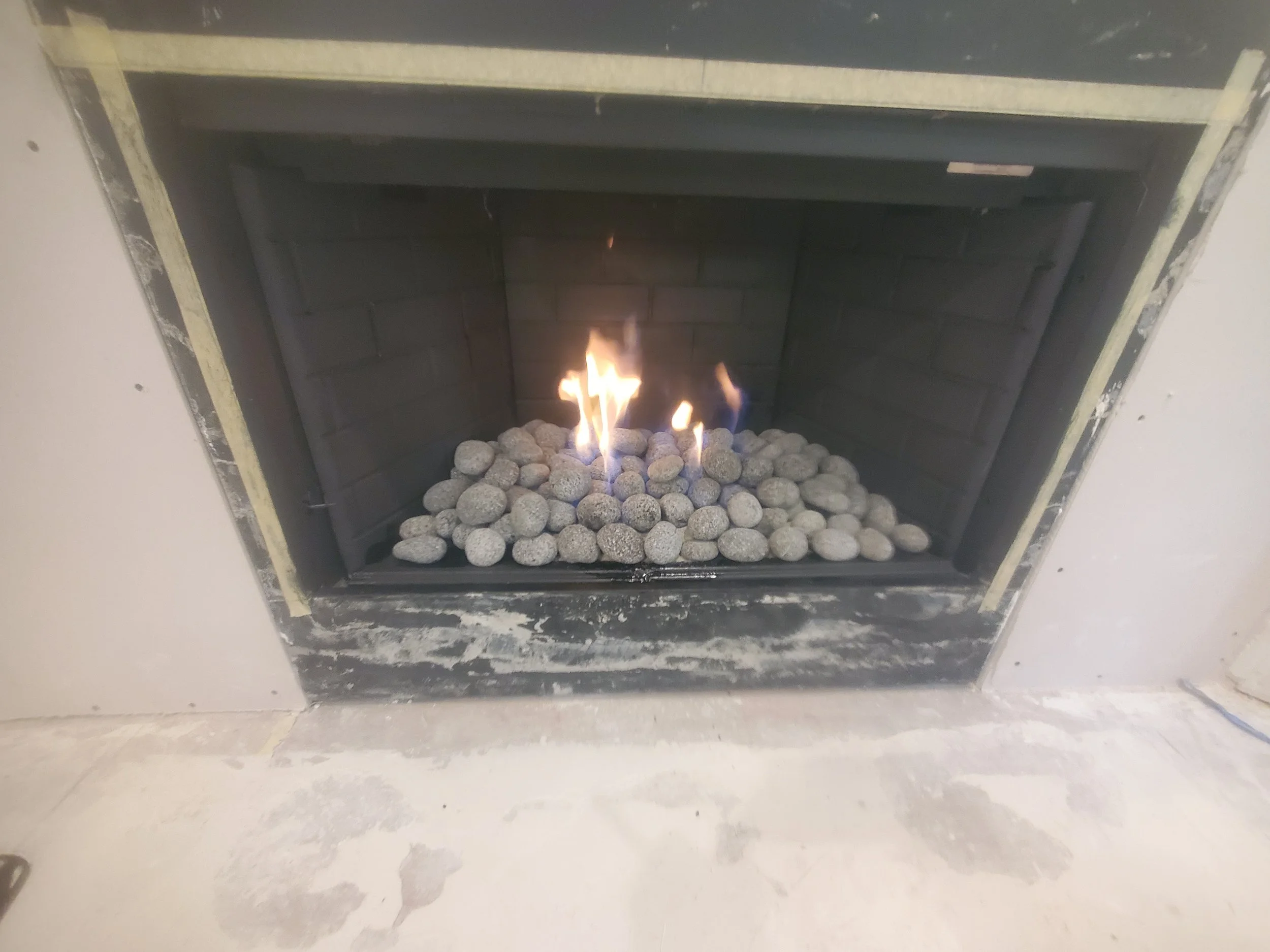 A fireplace with a small fire burning inside, surrounded by small gray stones. The fireplace is partially finished with an unfinished wall around it.
