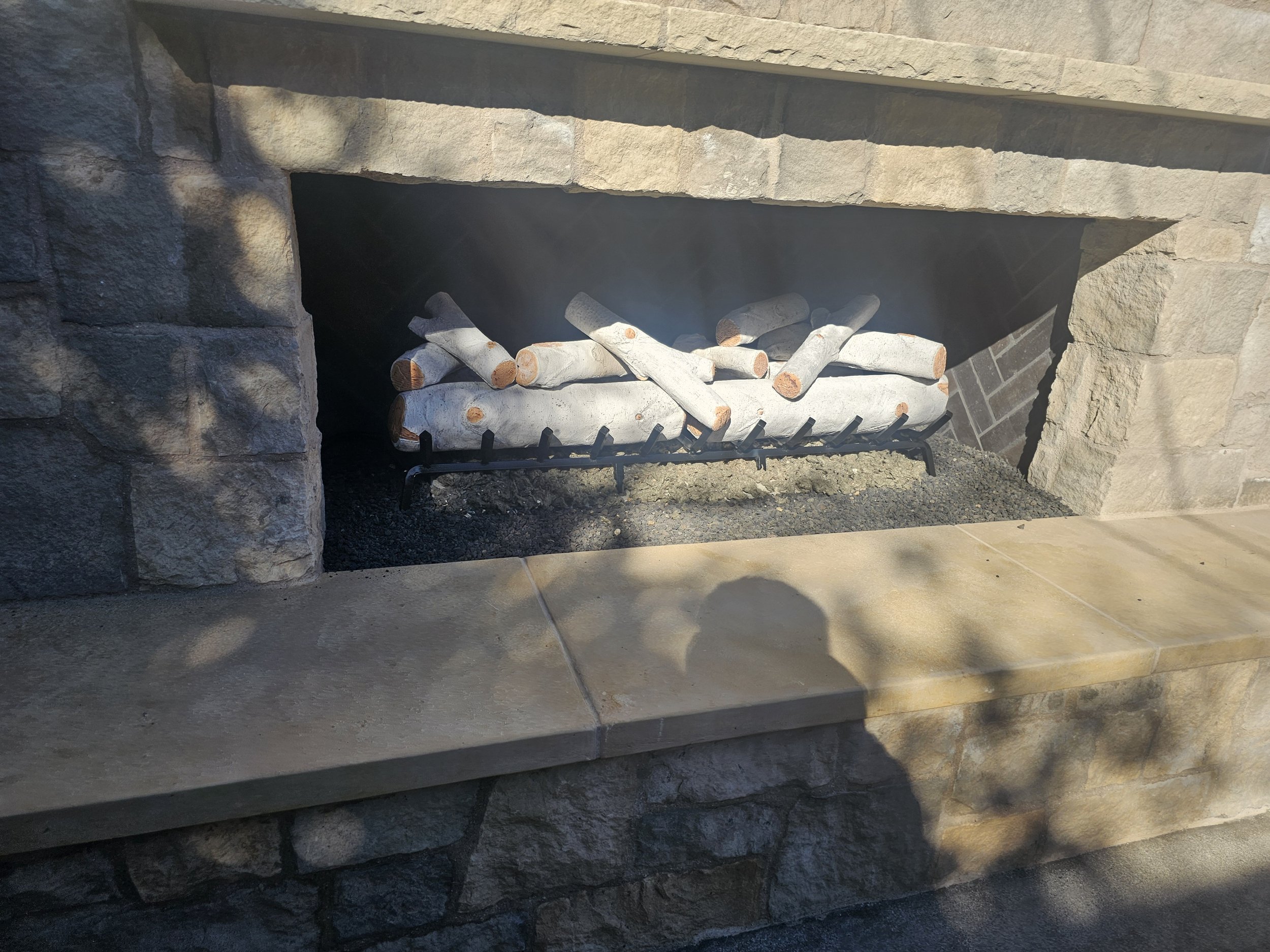 Logs of wood stored in a metal grate inside a stone fireplace. The fireplace has a stone mantle and surrounding stonework.