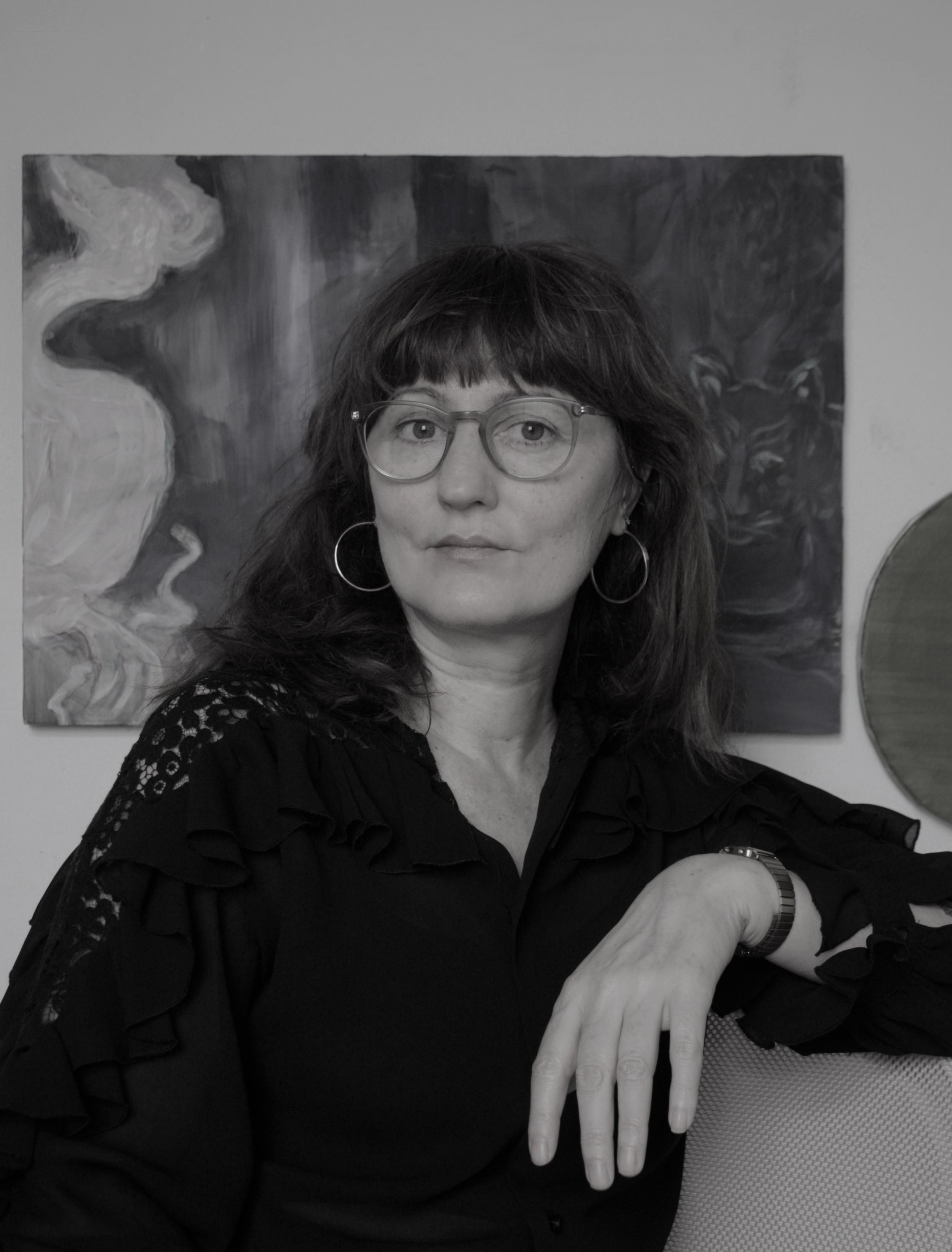 A woman with dark hair, glasses, and hoop earrings sitting in front of an abstract painting, wearing a black top with lace details on the shoulders.