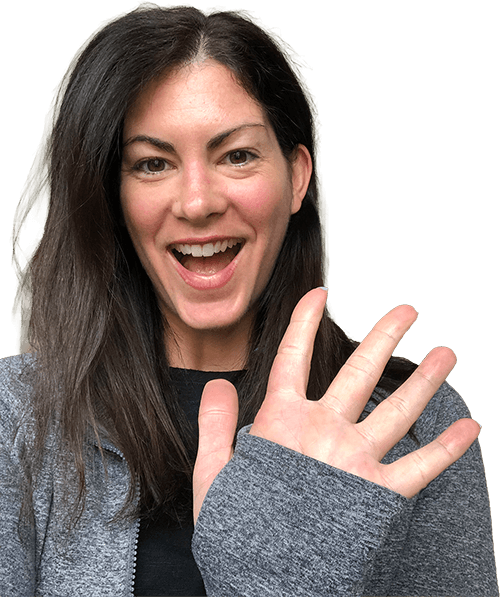 A woman smiling with an open mouth, waving with her right hand, wearing a gray jacket and black shirt.