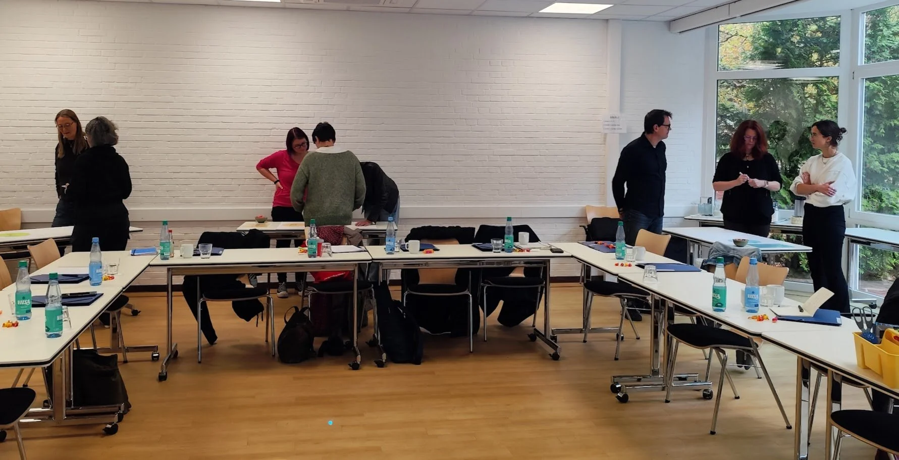 Gruppengespräch in einem Tagungsraum mit Tischen, Wasserflaschen, Notizbüchern und Stiften, Fenster mit Blick auf Bäume im Hintergrund.