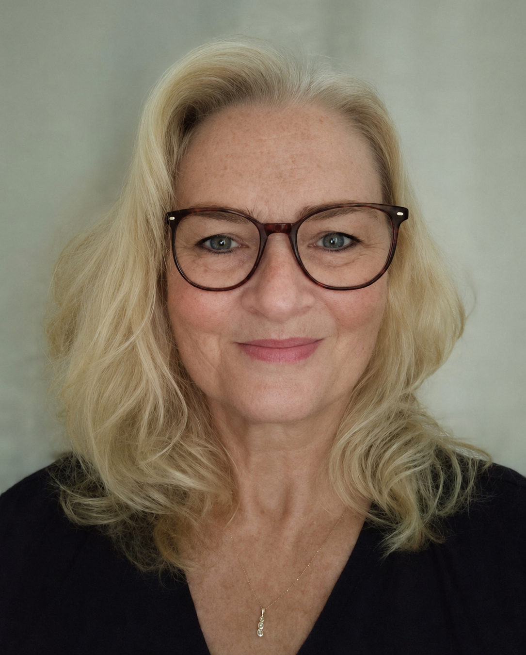 Nahaufnahme eines lächelnden Porträts einer Frau mit blonden Haaren, blauen Augen und Sommersprossen, die eine dunkle Jacke und ein blau-musterndes Halstuch trägt, vor einem neutralen Hintergrund.