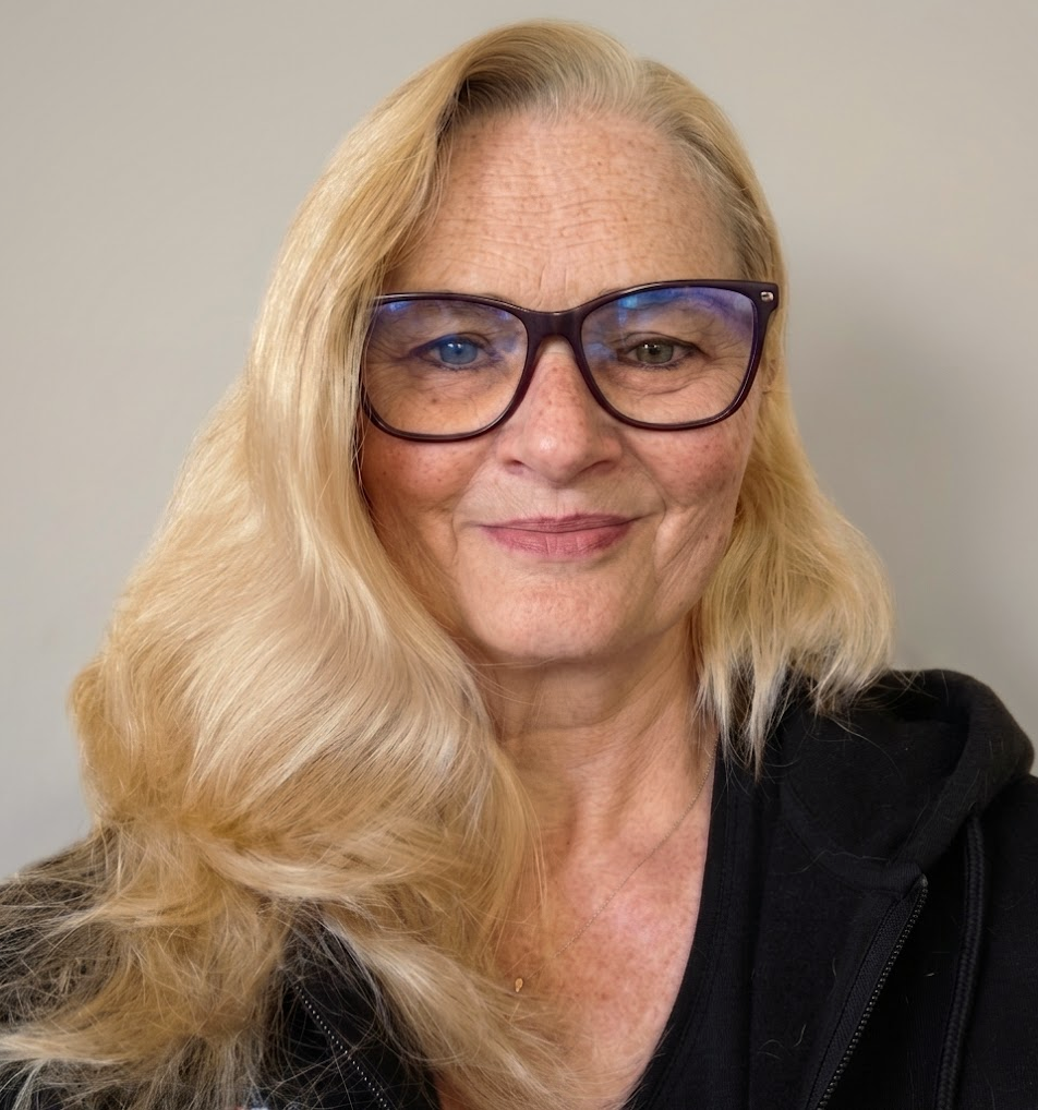 Nahaufnahme eines lächelnden Porträts einer Frau mit blonden Haaren, blauen Augen und Sommersprossen, die eine dunkle Jacke und ein blau-musterndes Halstuch trägt, vor einem neutralen Hintergrund.