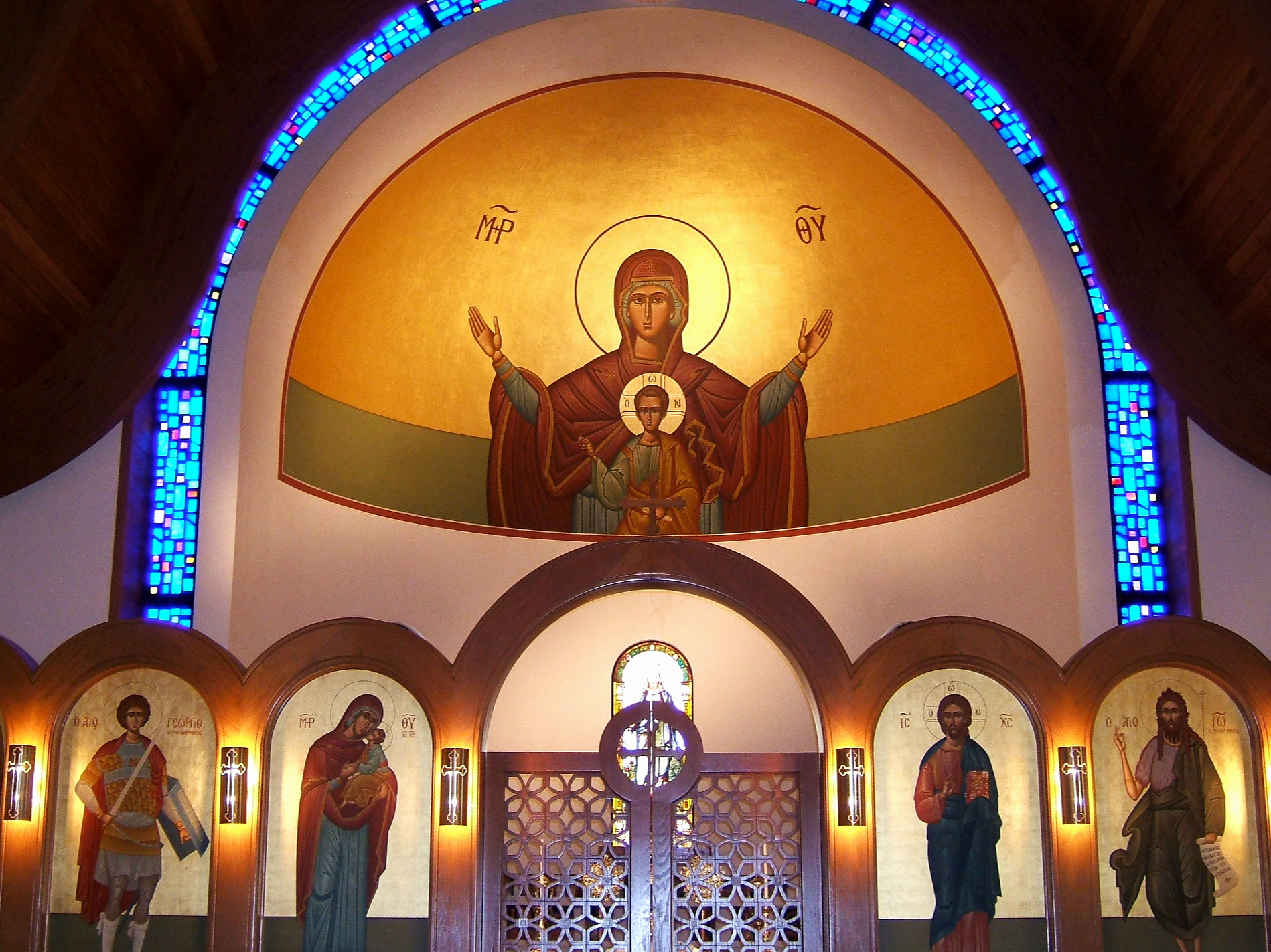 Inside view of St. George Greek Orthodox Church in OKC, featuring a large icon of the Virgin Mary with Christ, surrounded by smaller icons of saints, with decorative stained glass windows and wooden architectural elements.
