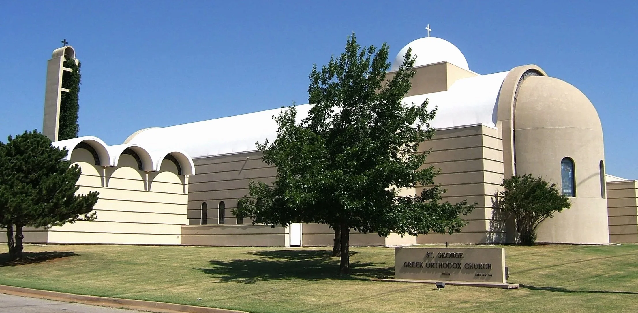 St. George Greek Orthodox church with a white dome and beige walls, surrounded by green trees and a grassy lawn, under a clear blue sky in Oklahoma City.