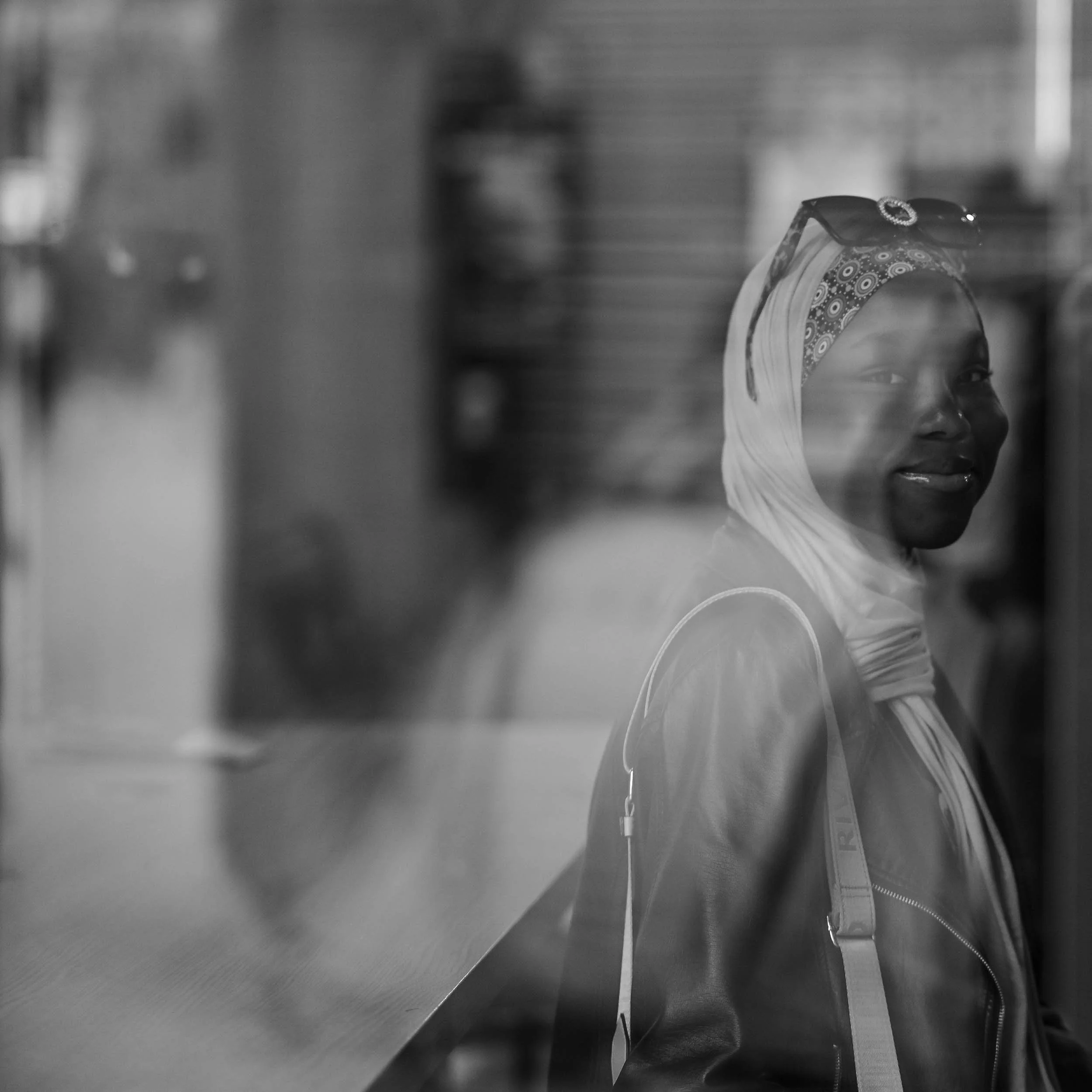 Black and white image showing a woman with a headscarf looking over a metallic counter or surface, possibly in a kitchen or work environment.