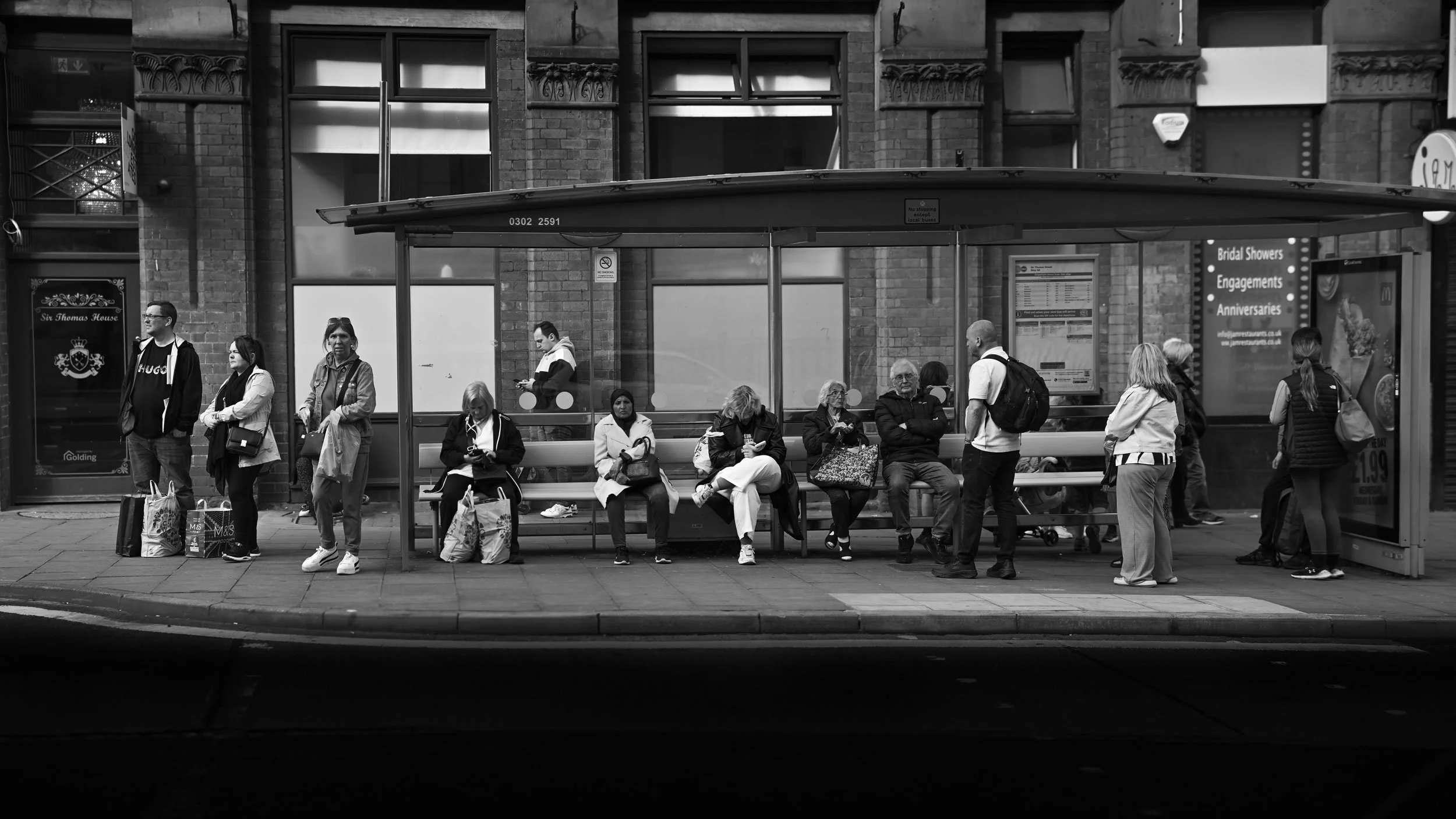 People waiting at a bus stop in an urban area with a stone building in the background. Some are seated, others are standing, carrying bags or looking at their phones.