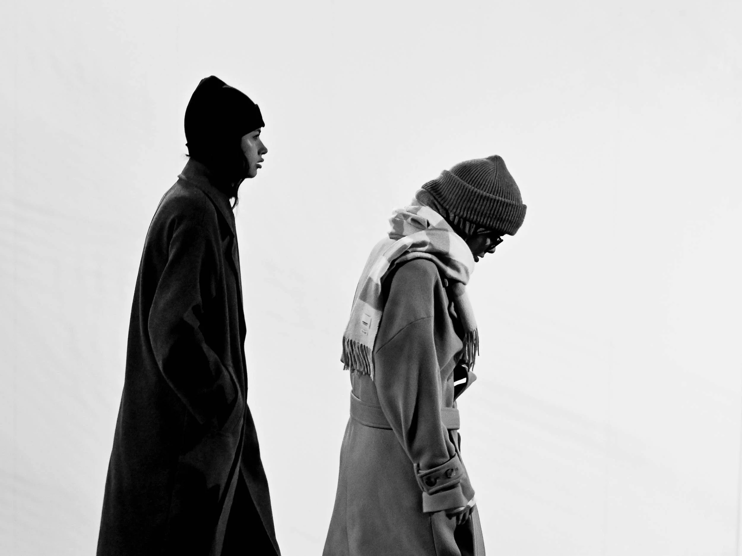 Side profile of a woman wearing a beanie and a man wearing glasses and a ribbed beanie, both dressed in warm clothing, walking outdoors against a plain sky.