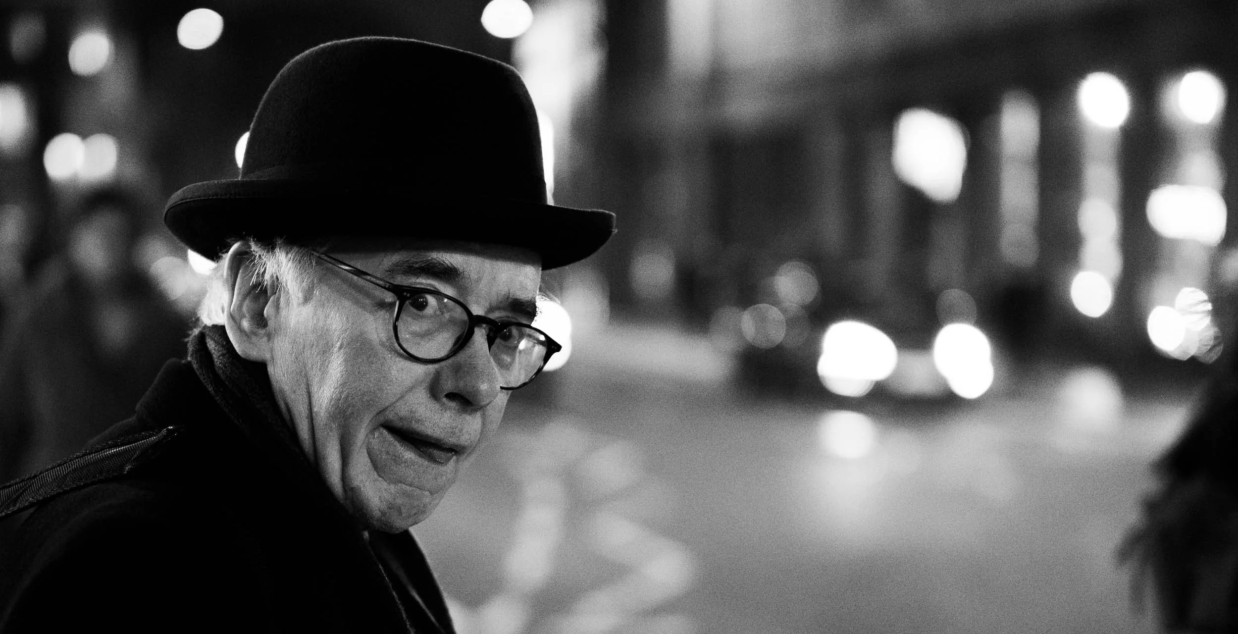 A black and white photo of an elderly man with glasses, wearing a fedora hat, smiling and looking sideways, with blurred city lights in the background at night.