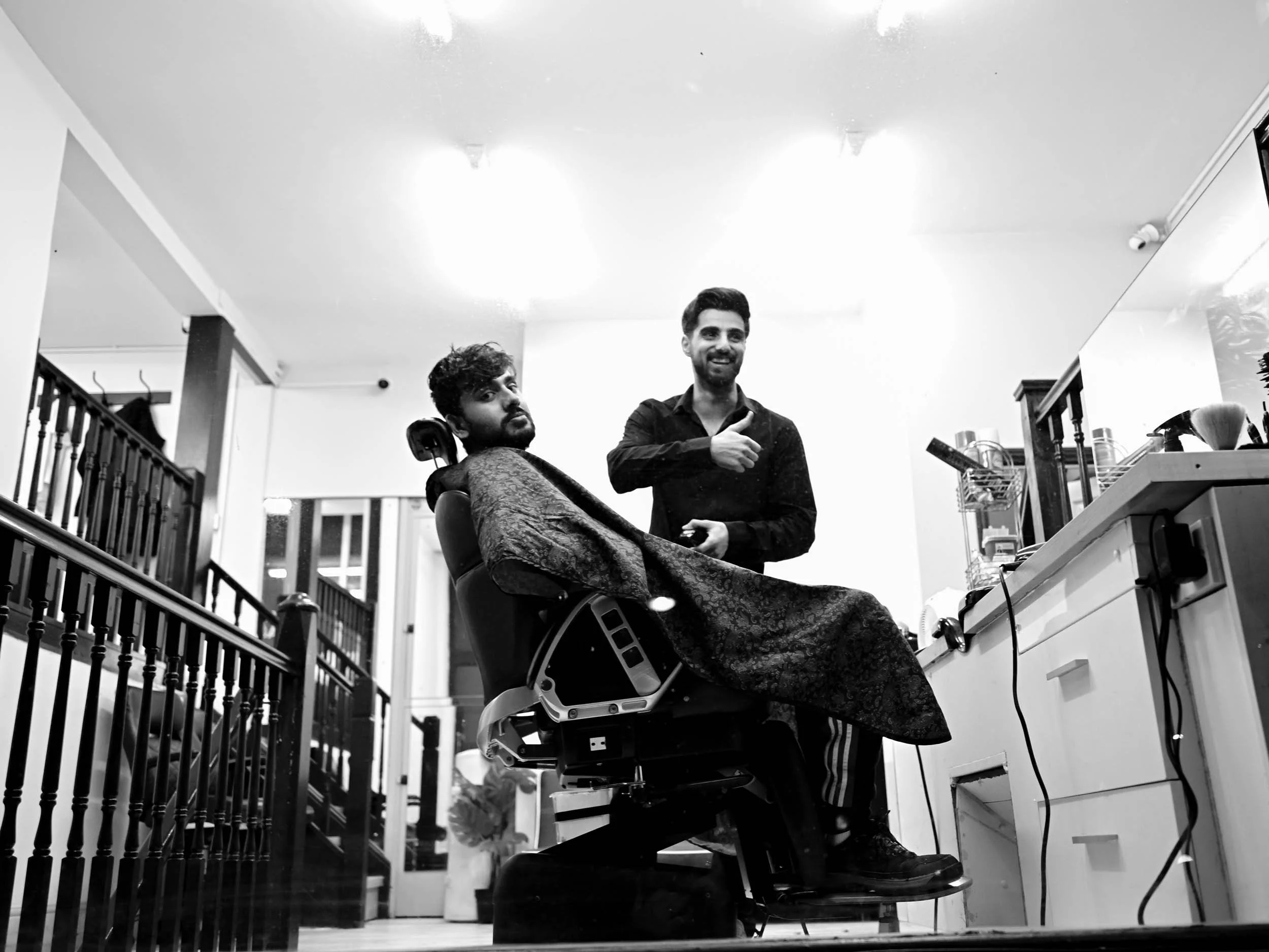 A man in a barber shop getting a haircut while another man, possibly the barber, smiles and gestures with his hand. The scene is in black and white.