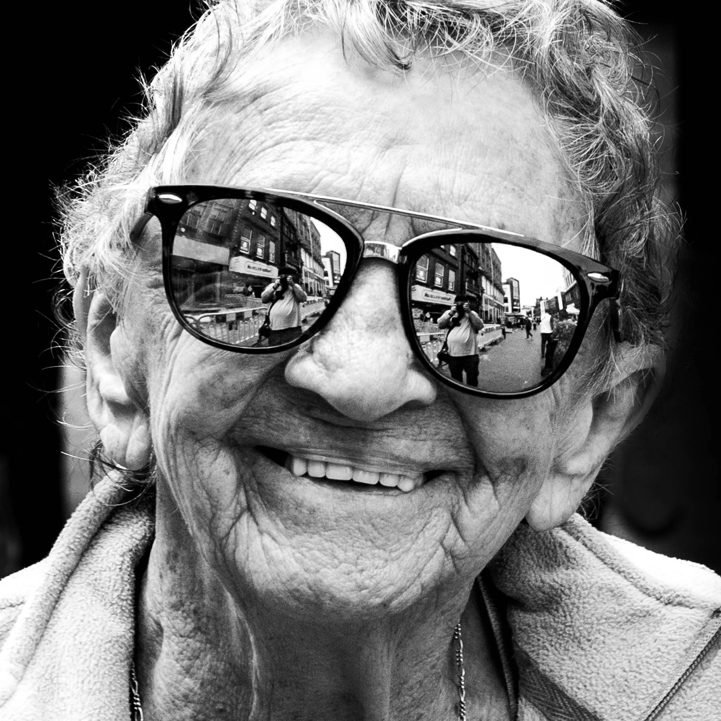 Close-up black and white photo of a smiling elderly woman with curly hair wearing dark sunglasses and a fleece jacket, with reflected buildings and photographer in the sunglasses.