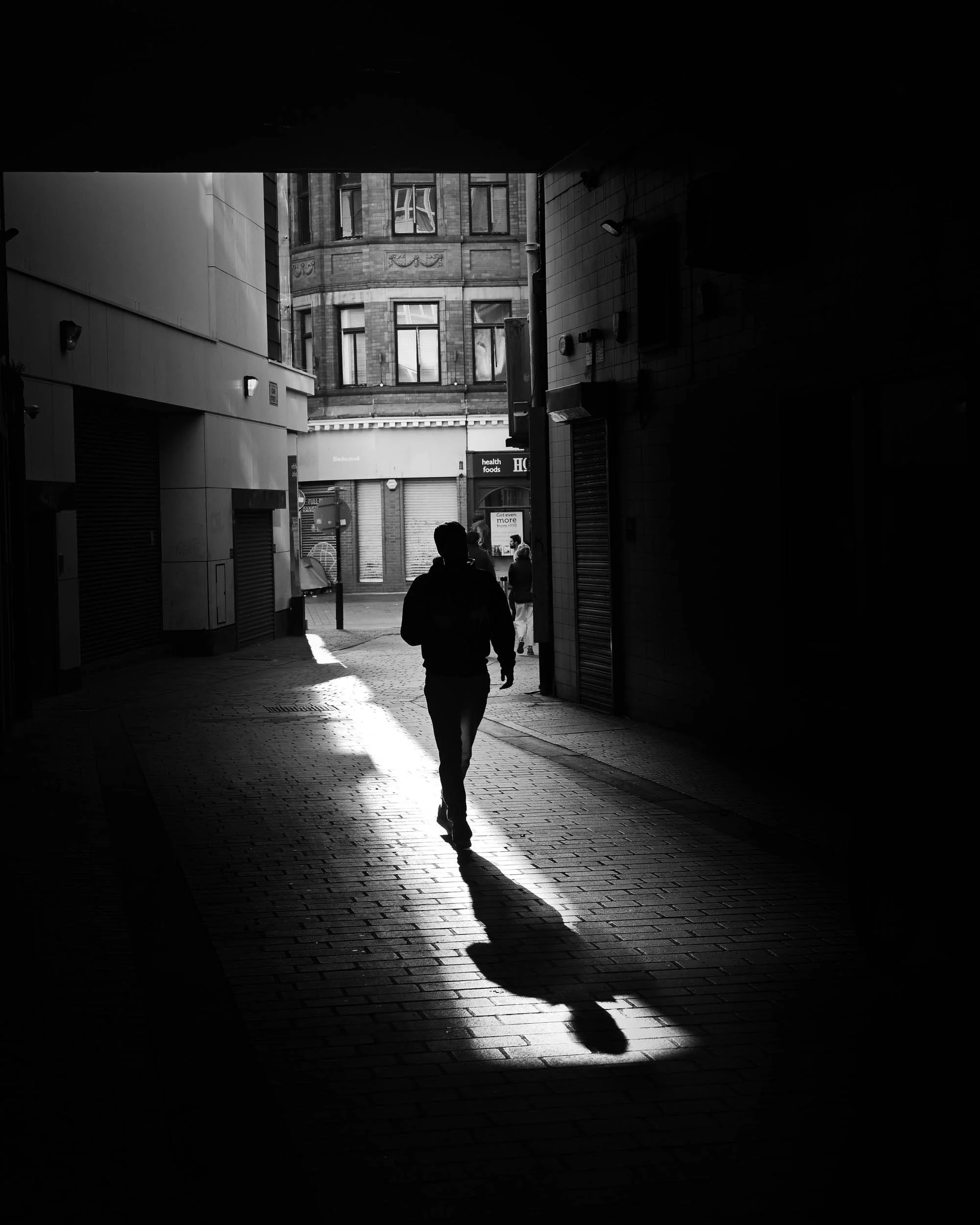 A silhouette of a person walking through a dark alleyway toward a brighter street, with sunlight illuminating the cobblestone pavement and the building facades in the background.