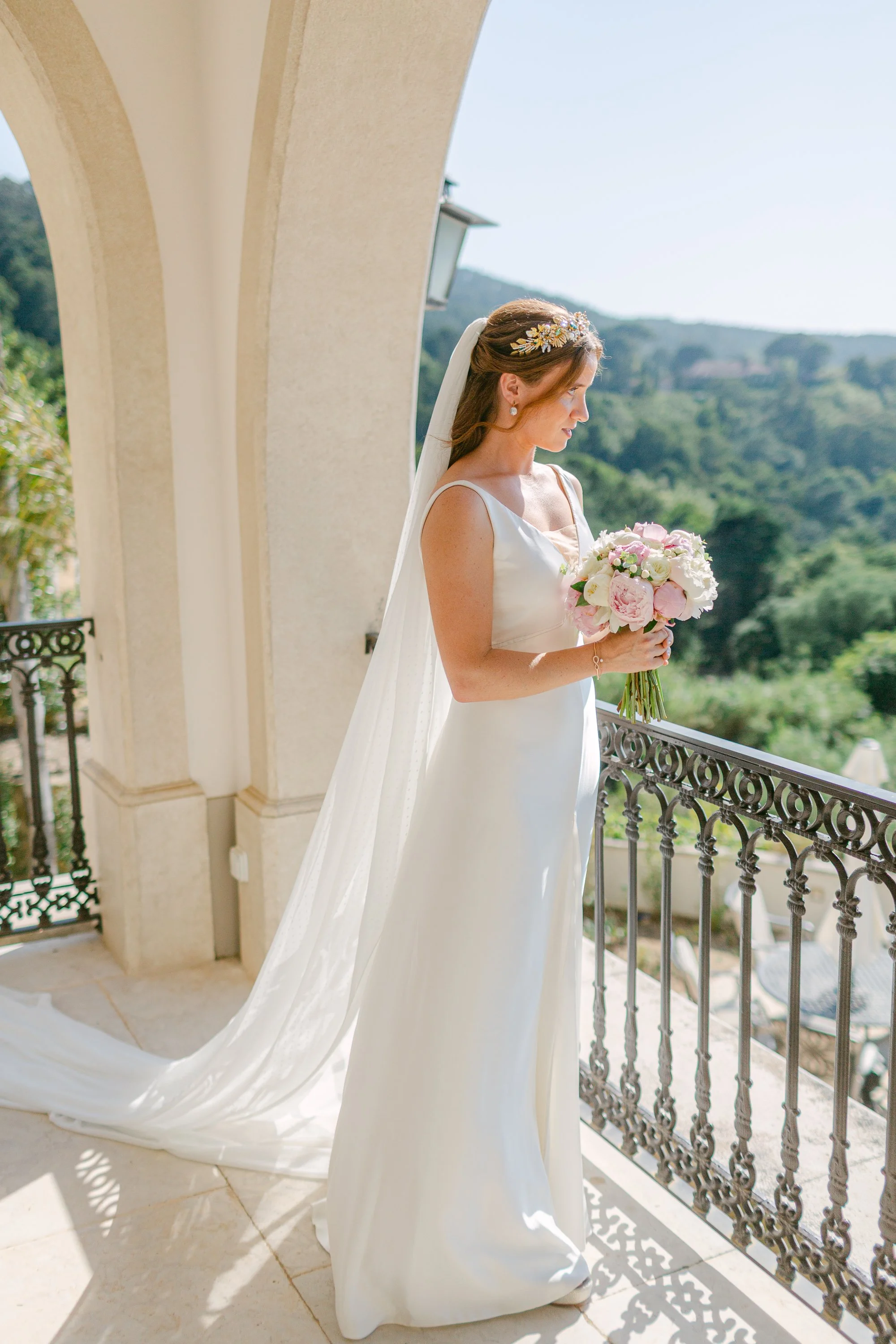Bride in white wedding dress holding a bouquet on a balcony with a scenic green landscape in the background.