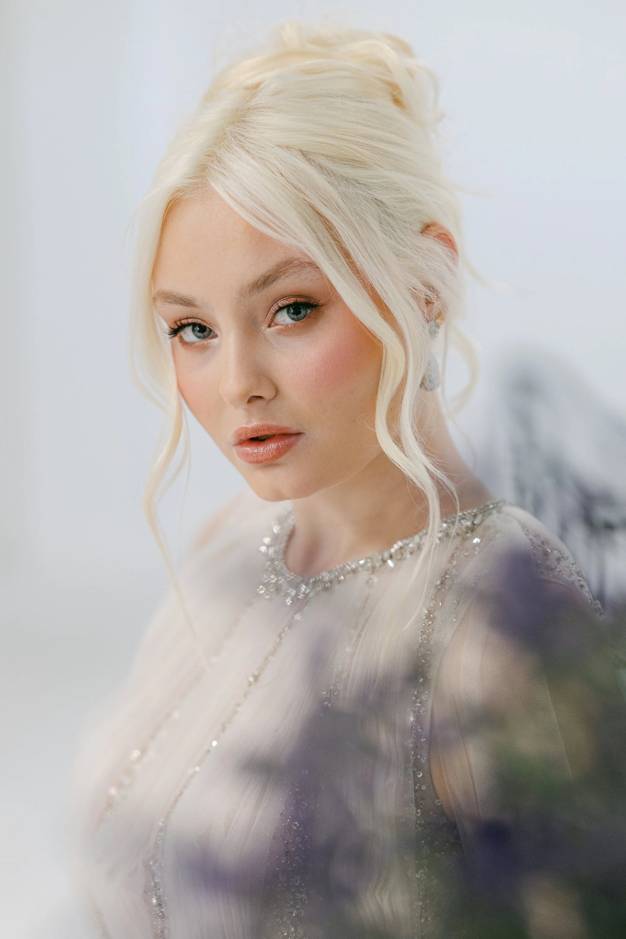 A woman with platinum blonde hair styled in loose waves, wearing a sparkly, sheer top with jewel accents, and looking directly at the camera against a plain, light background.