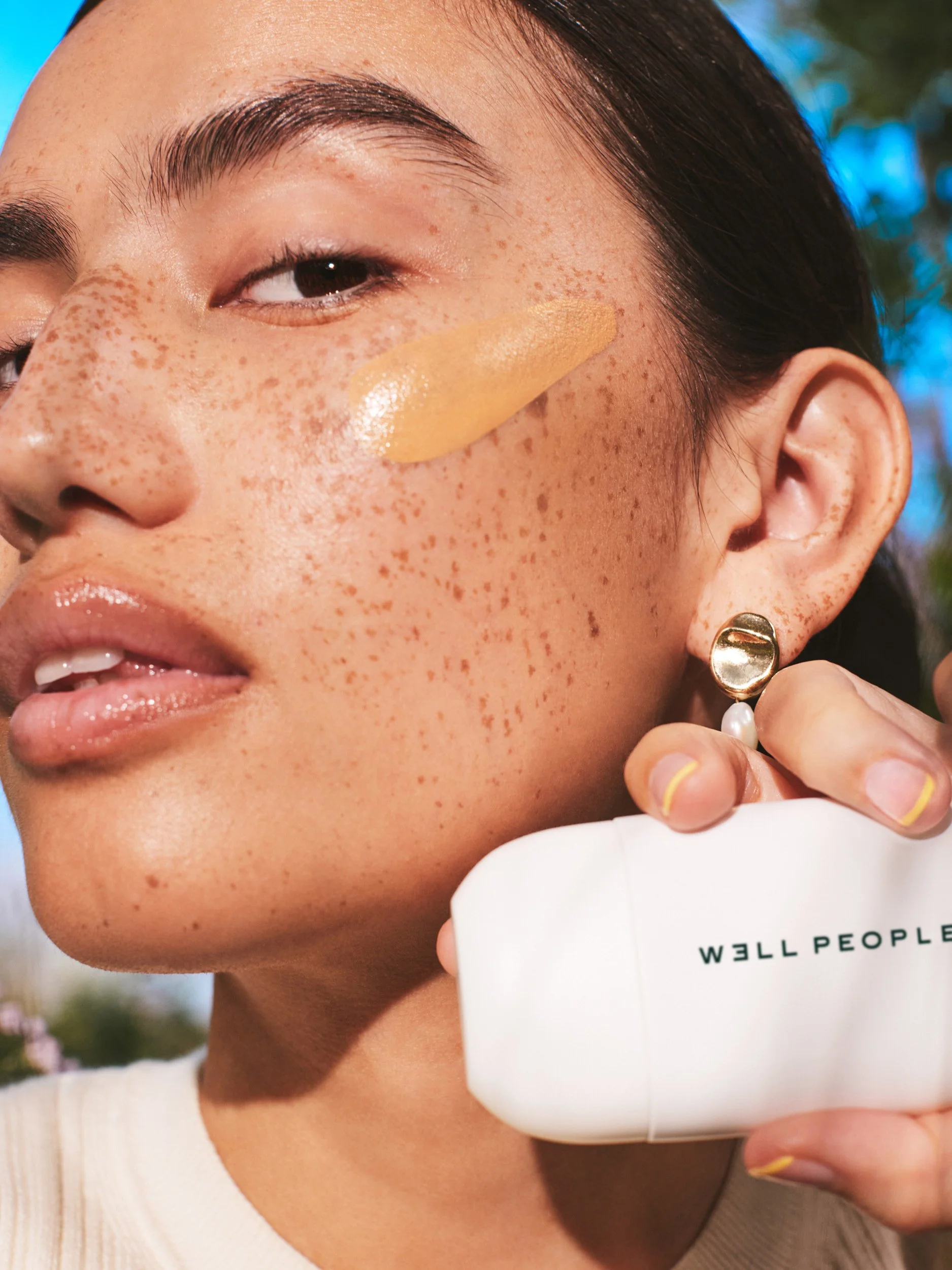 Joshua Pestka commercial beauty photography — Well People Skinphoria campaign, model with freckled skin holding Well People compact, product swatch on cheek, close-up
