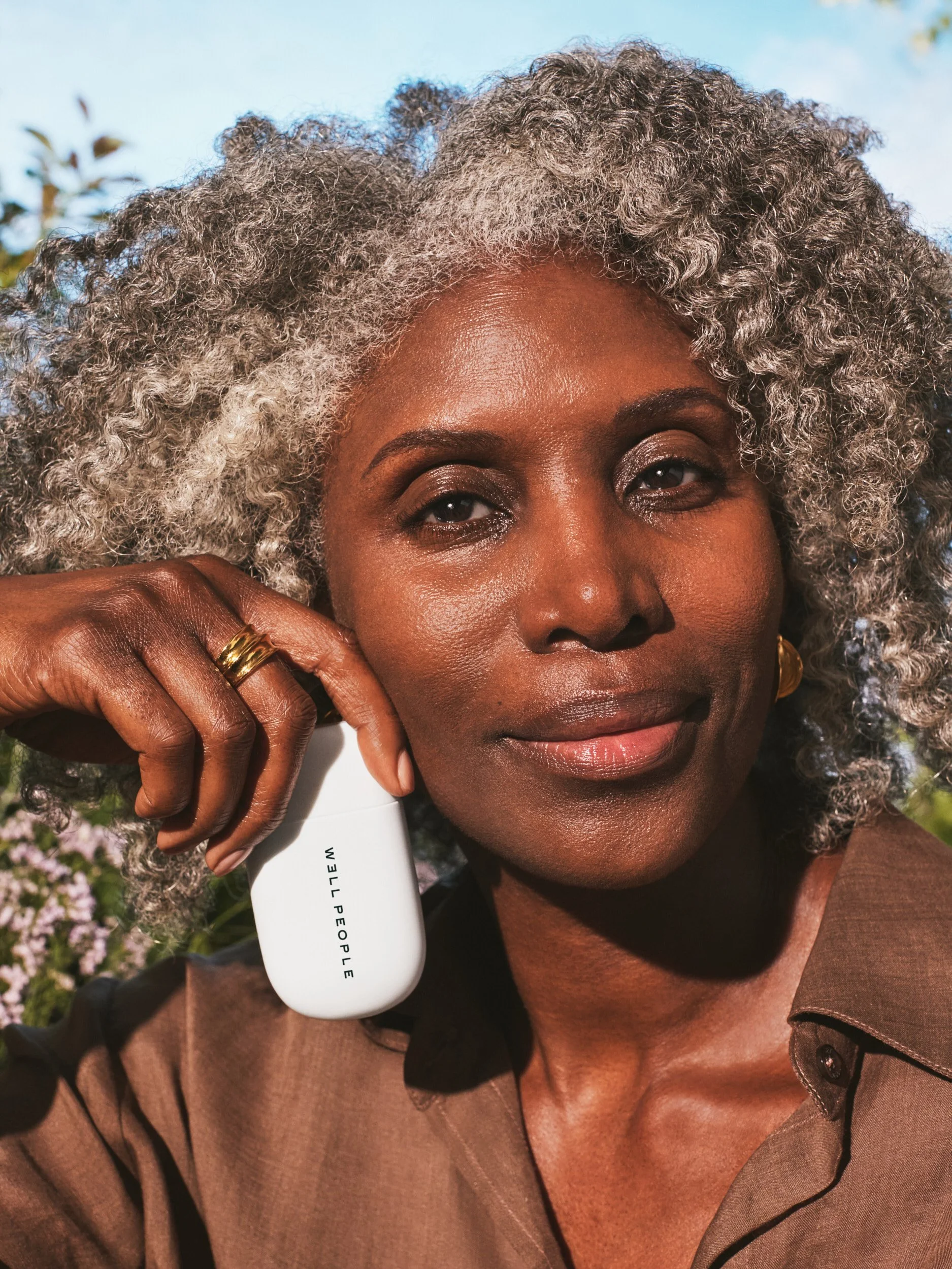 Joshua Pestka commercial beauty photography — Well People Skinphoria campaign, model with silver afro holding Well People product to cheek, warm outdoor light