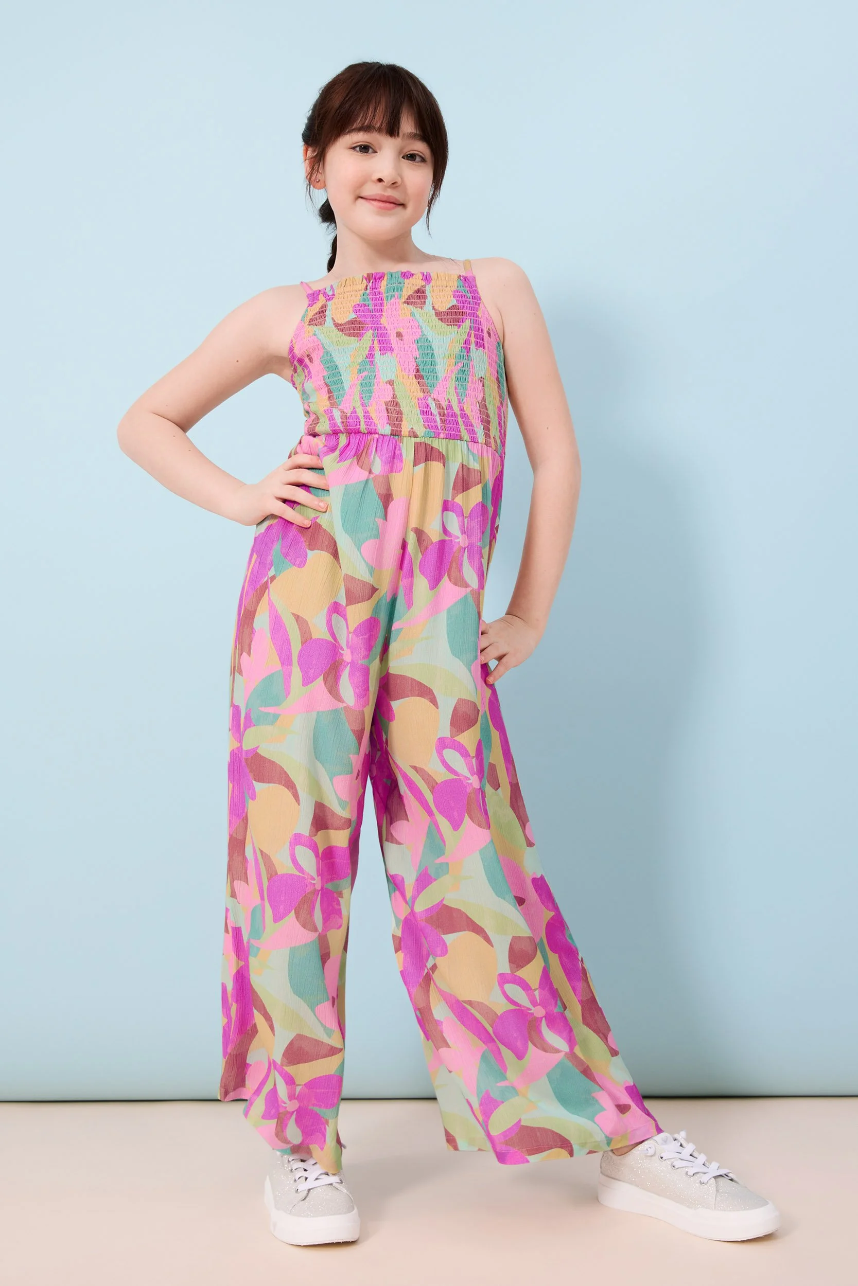 Joshua Pestka kids fashion photography — girl in floral wide-leg jumpsuit and white sneakers, standing pose, light blue background