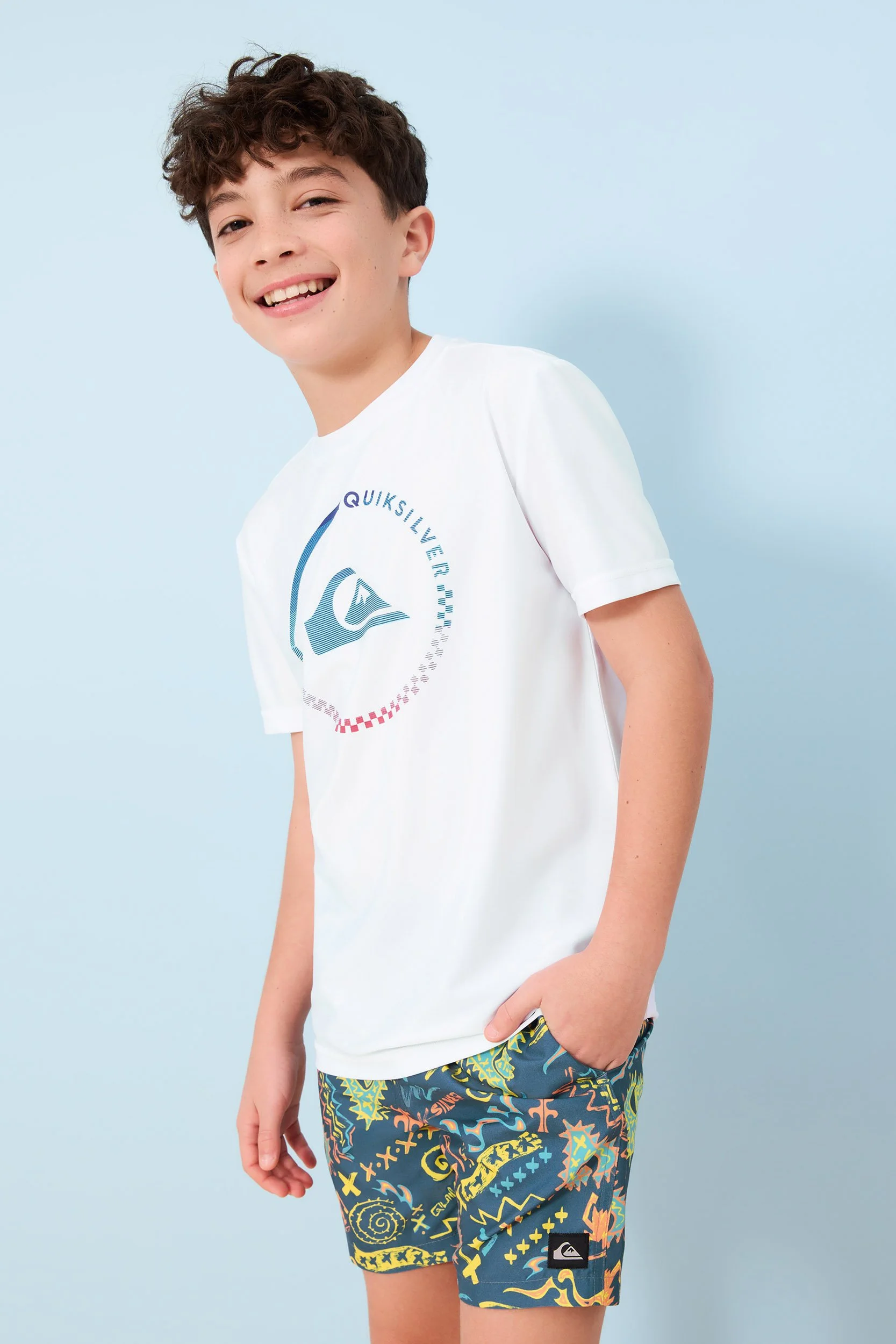 Joshua Pestka kids fashion photography — boy in Quiksilver white graphic tee and printed shorts, standing pose, light blue background