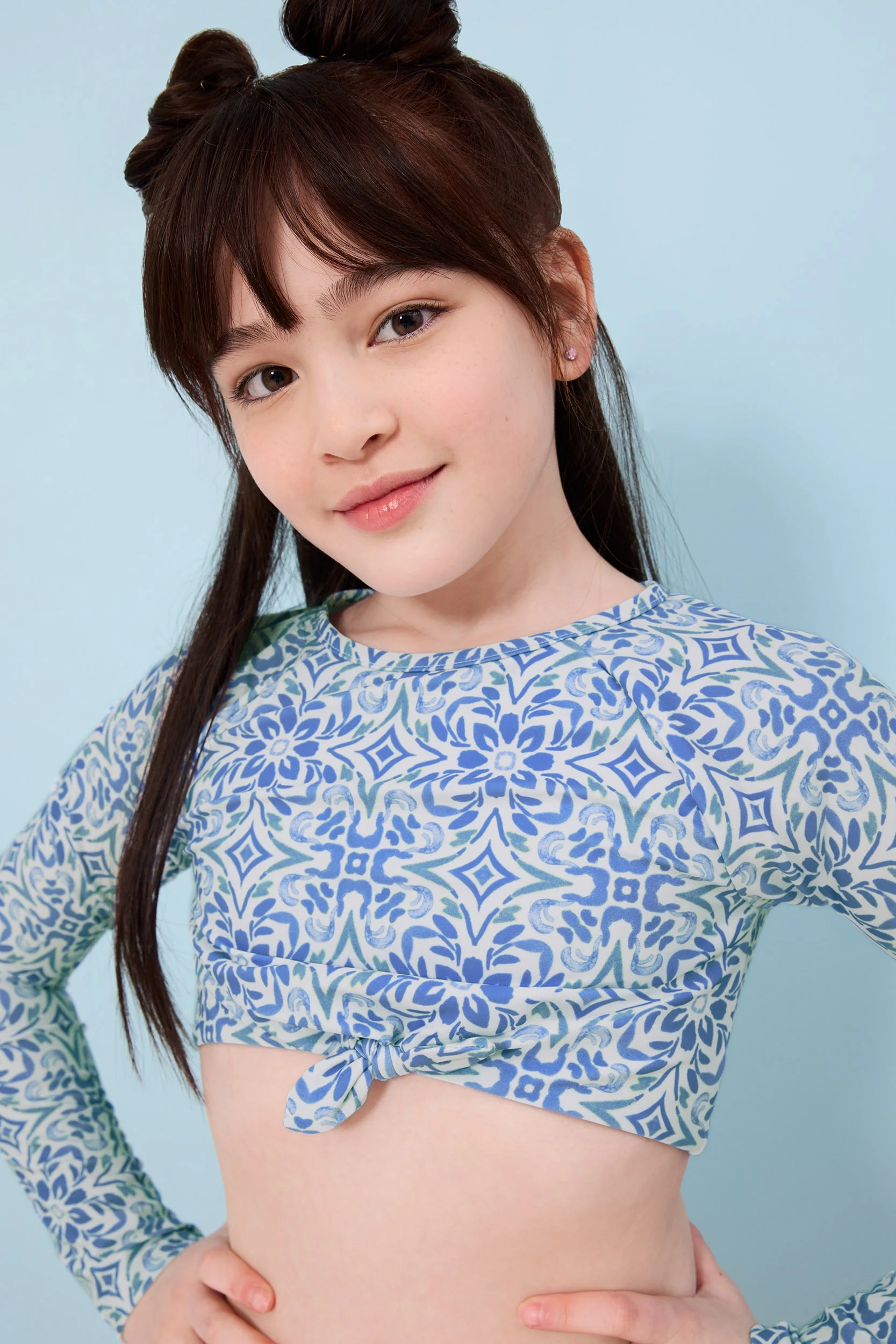Joshua Pestka kids fashion photography — girl in blue floral crop top, dark hair with buns, direct gaze, white background