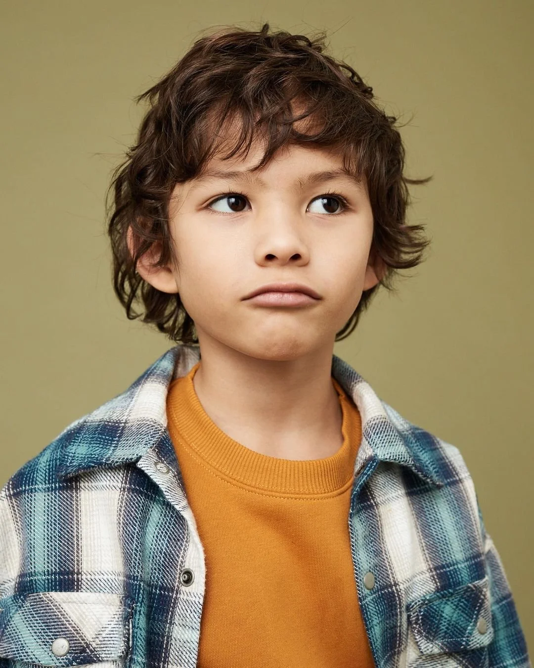 Joshua Pestka kids fashion photography — boy in plaid flannel shirt and mustard sweater, close-up portrait, olive background