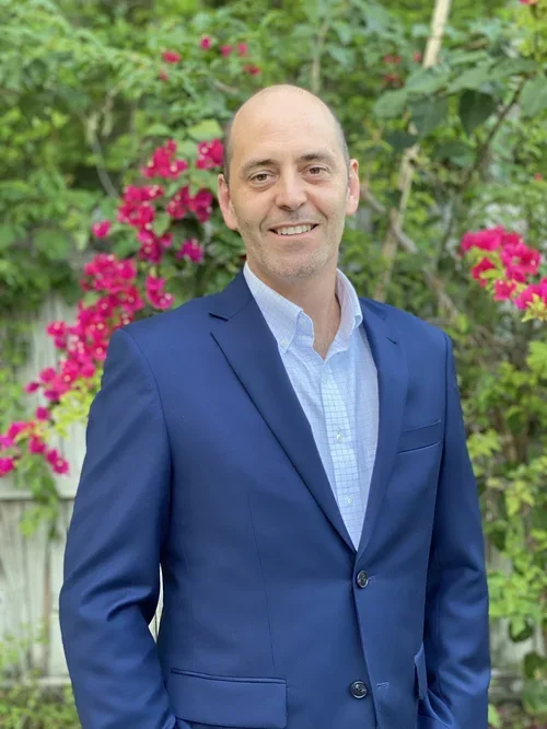 A man in a blue suit and light blue shirt standing outdoors with pink flowers and green foliage in the background.