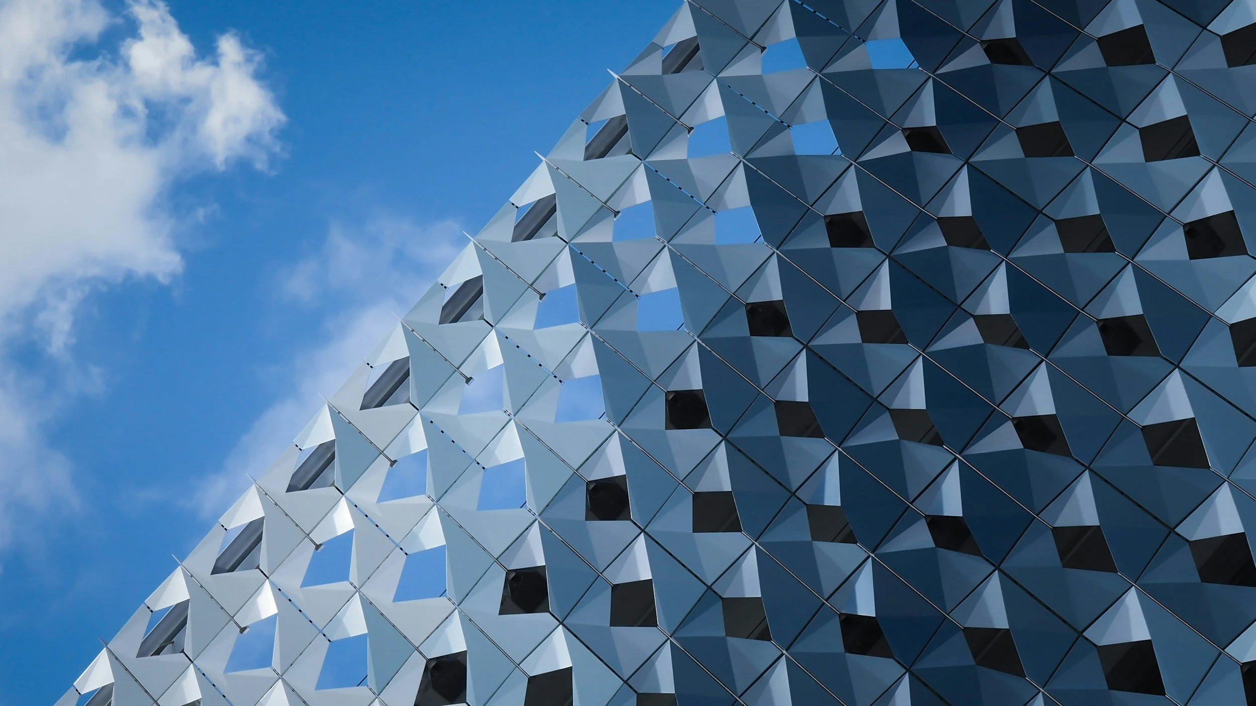 Close-up view of a modern building with a geometric patterned facade made of reflective metallic panels, against a bright blue sky with scattered clouds.