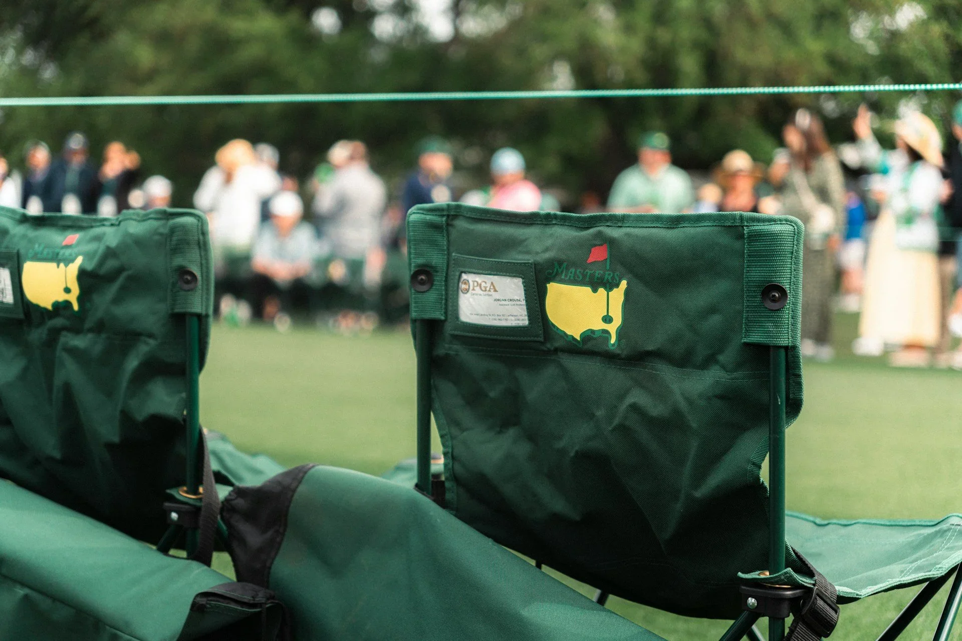 Two green chairs with a map of the United States and the PGA logo on the backrests, set on a grassy area with a crowd of people in the background.