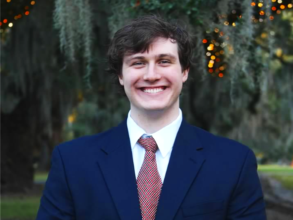 A young man in a navy blue suit, white shirt, and patterned tie, smiling outdoors with trees and holiday lights in the background.