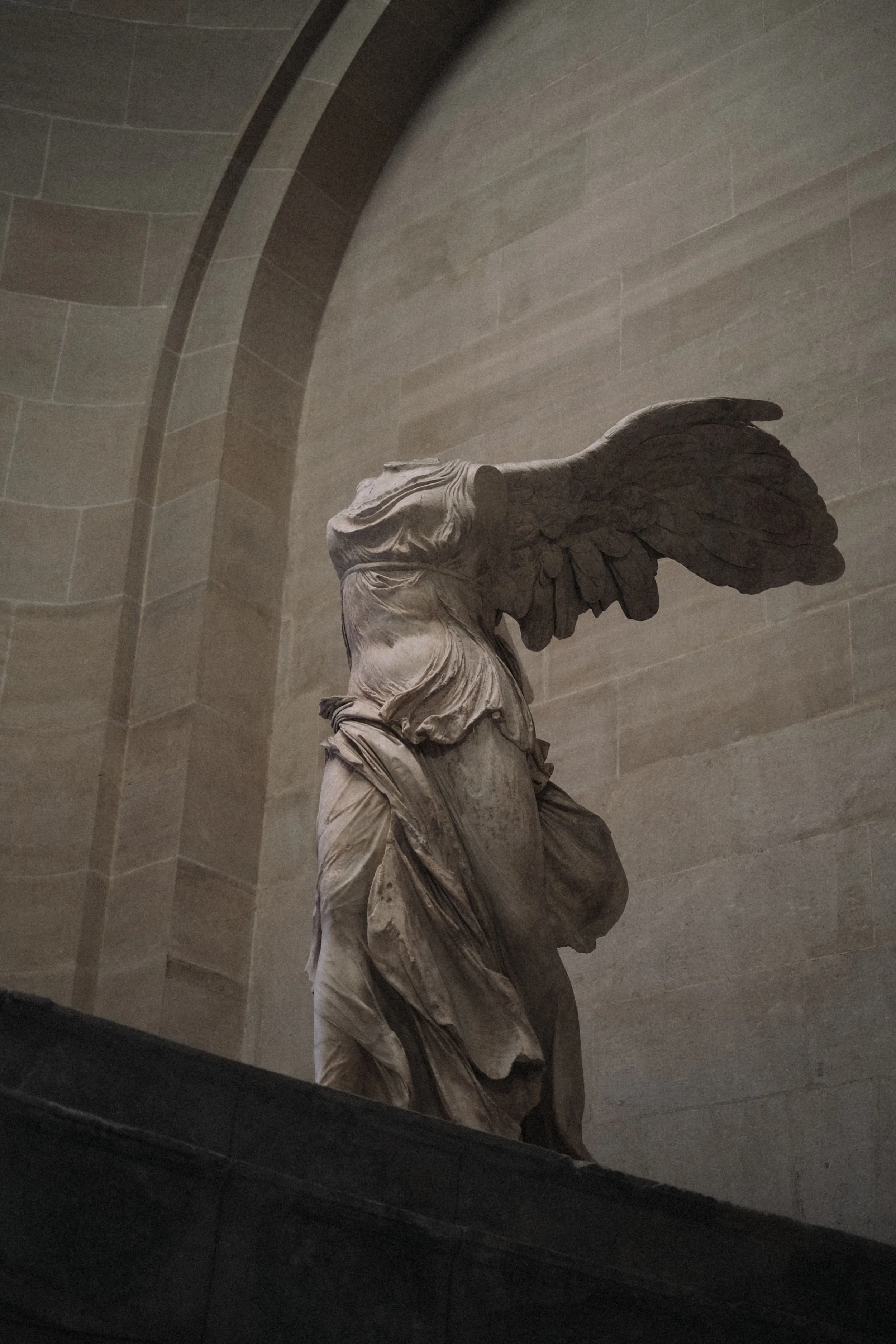 A marble statue of Nike, the Greek goddess of victory, with large wings, located inside a building with stone walls and an arched ceiling.