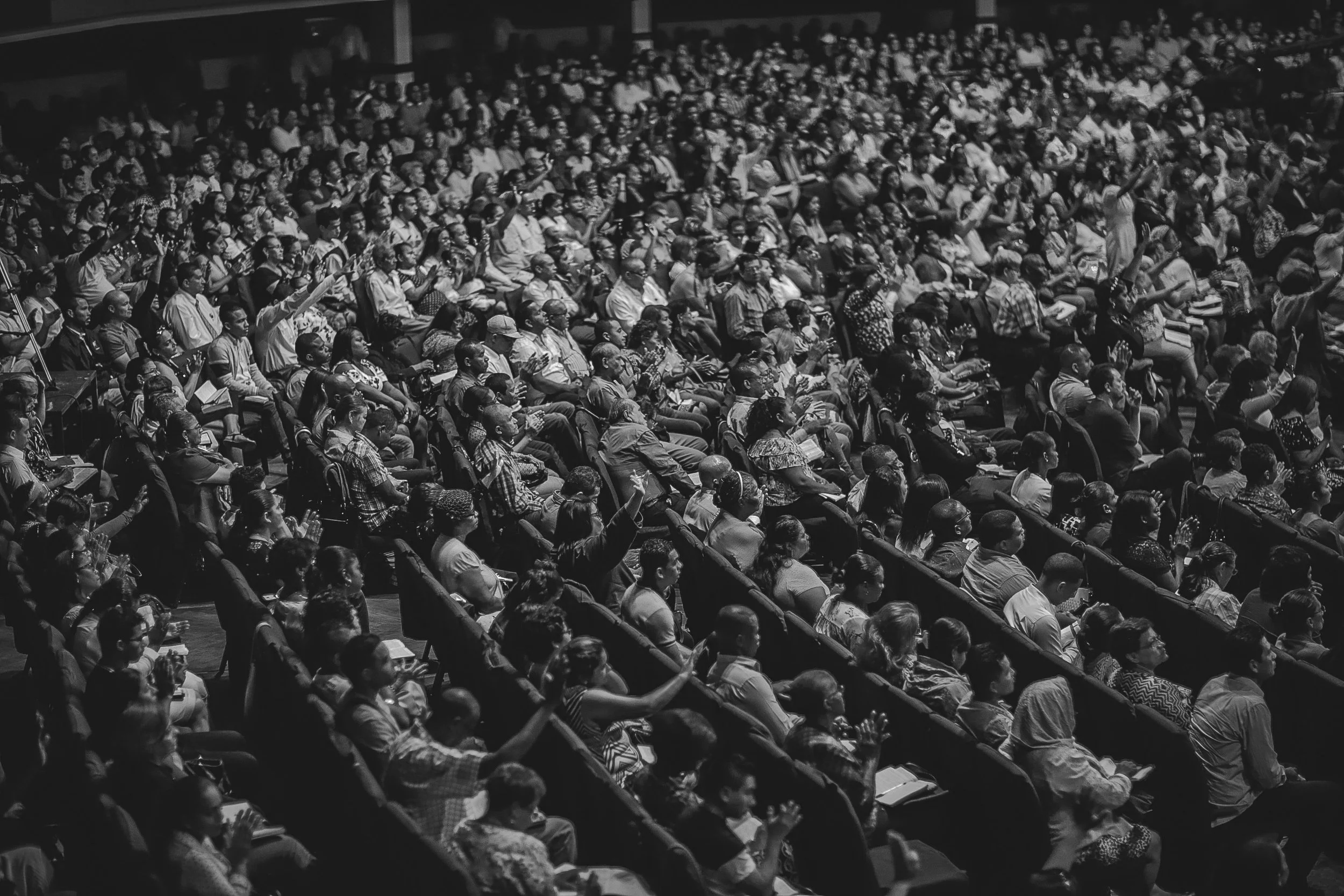 Black and white photo of a large audience attending a conference or event, seated in rows with some raising their hands.