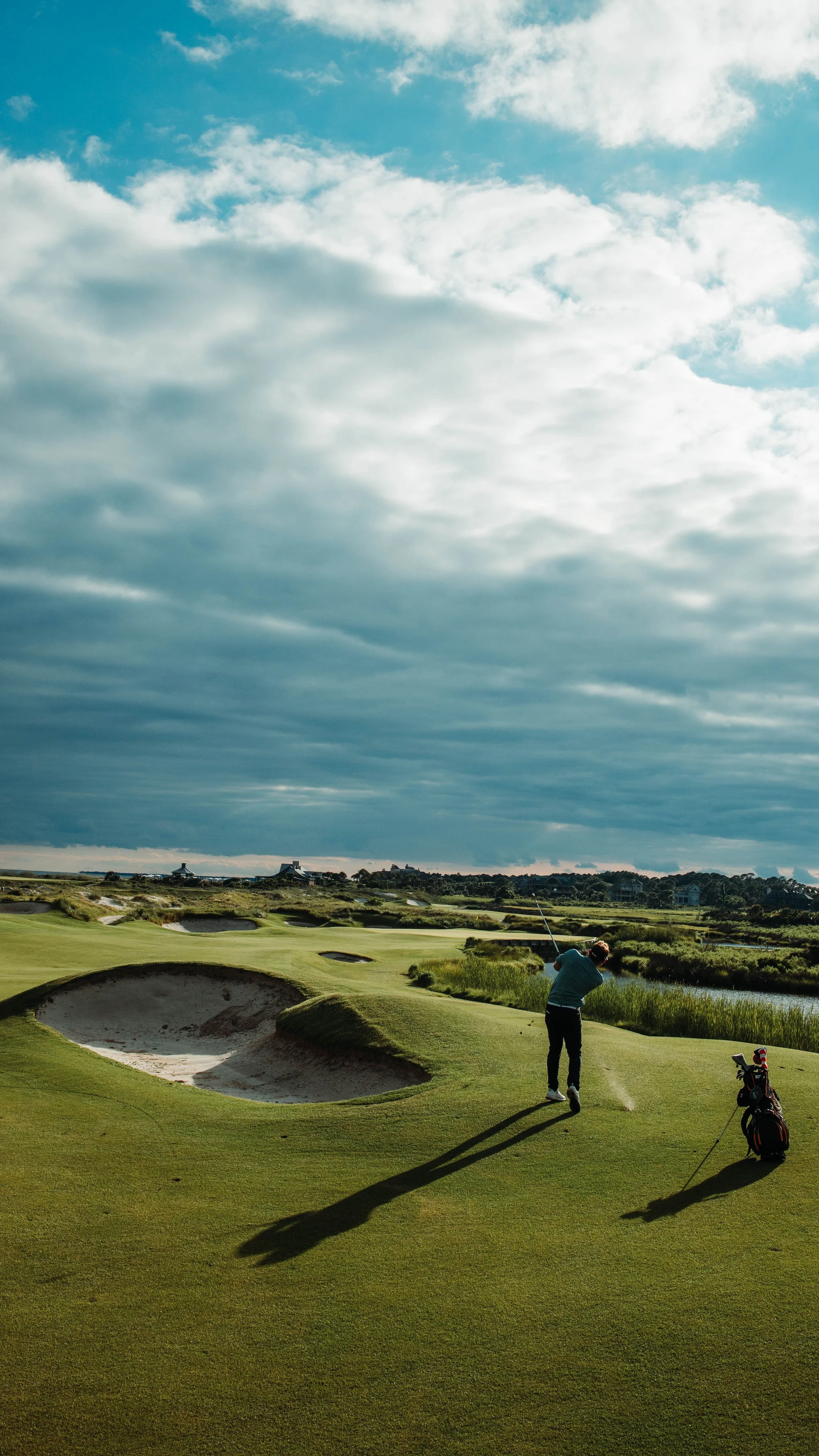 A golfer taking a shot on a golf course with a basket of golf clubs nearby, surrounded by sand traps, grass, and a partly cloudy sky.