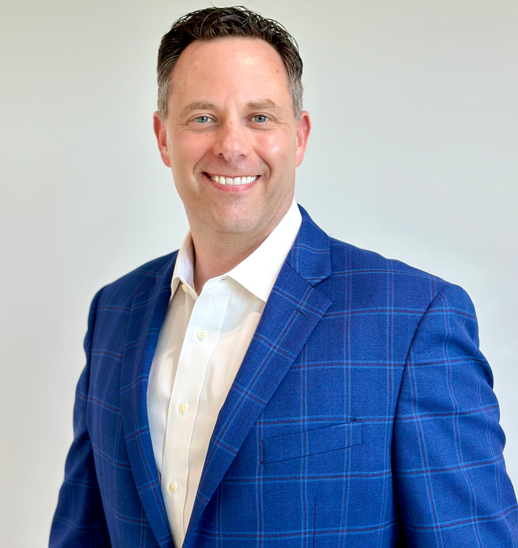 A smiling man wearing a blue plaid suit and a white shirt, posing against a plain light-colored background.