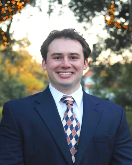 Young man in a suit and tie smiling outdoors with trees and sunlight in the background.