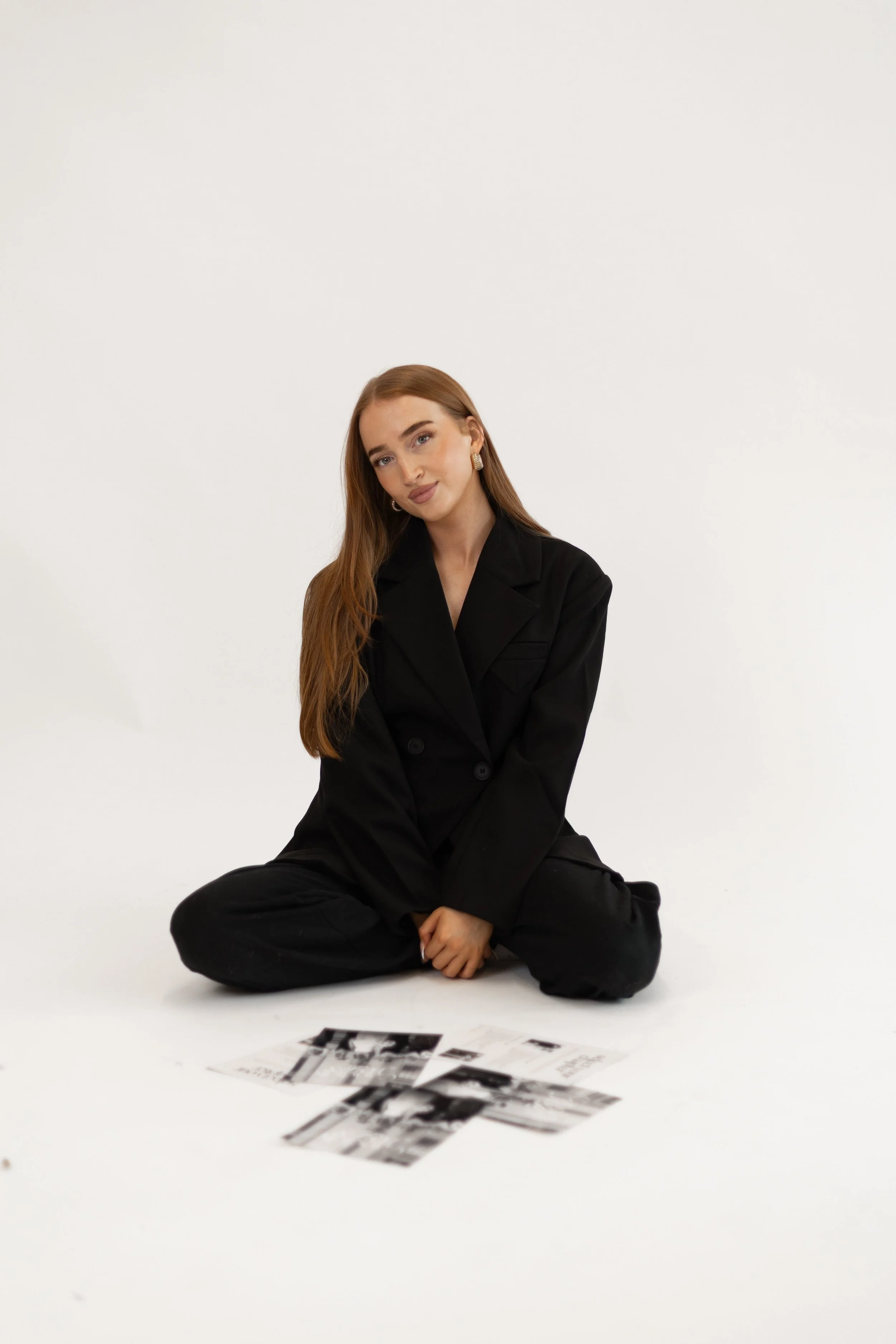 A young woman with long red hair sitting cross-legged on the floor, wearing a black blazer and pants, with photographs spread in front of her on a white background.