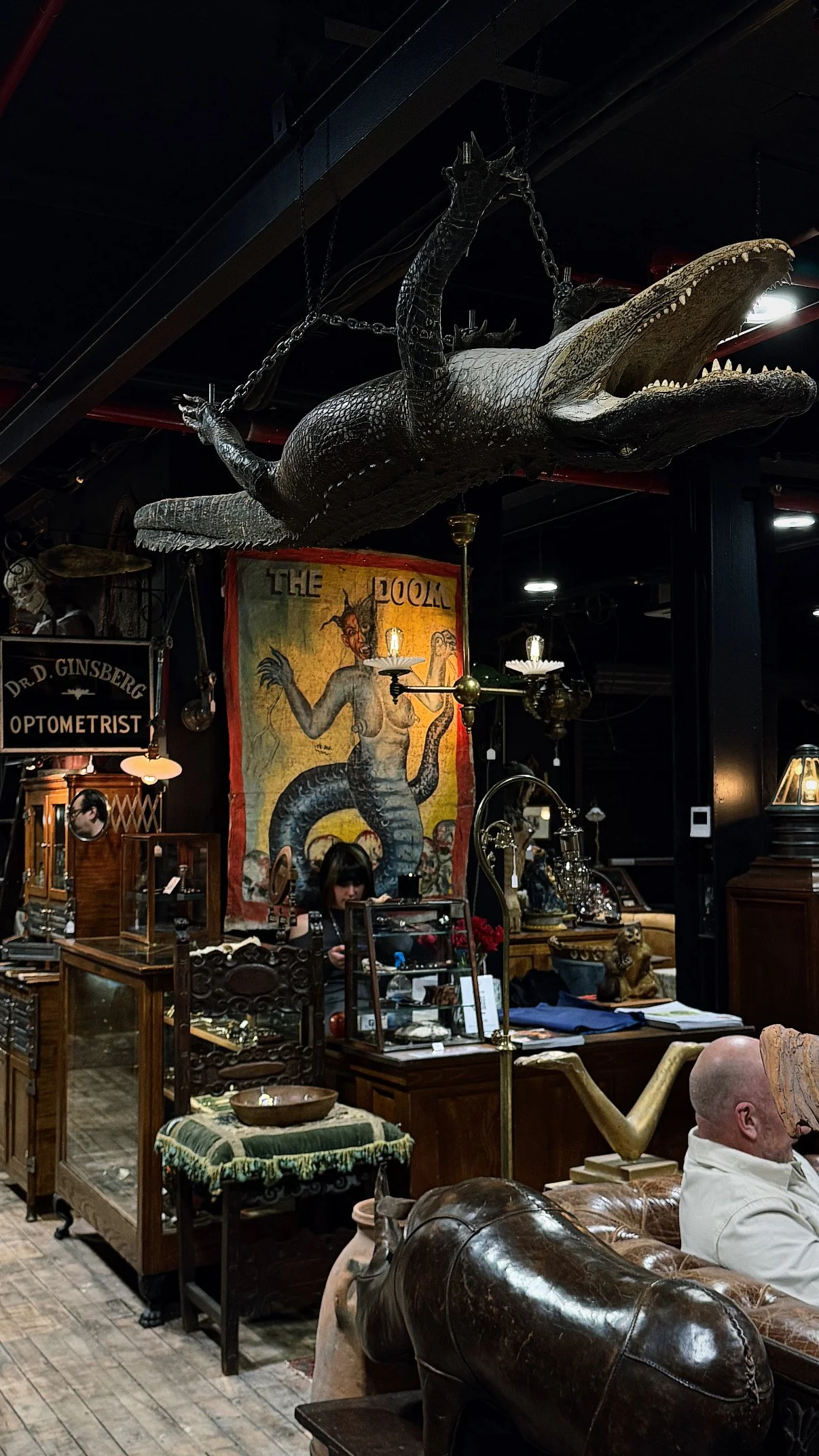 A vintage store filled with antique furniture and artwork, featuring a large, hanging crocodile sculpture above a lady working behind a counter, with a colorful poster of a mythical creature in the background.