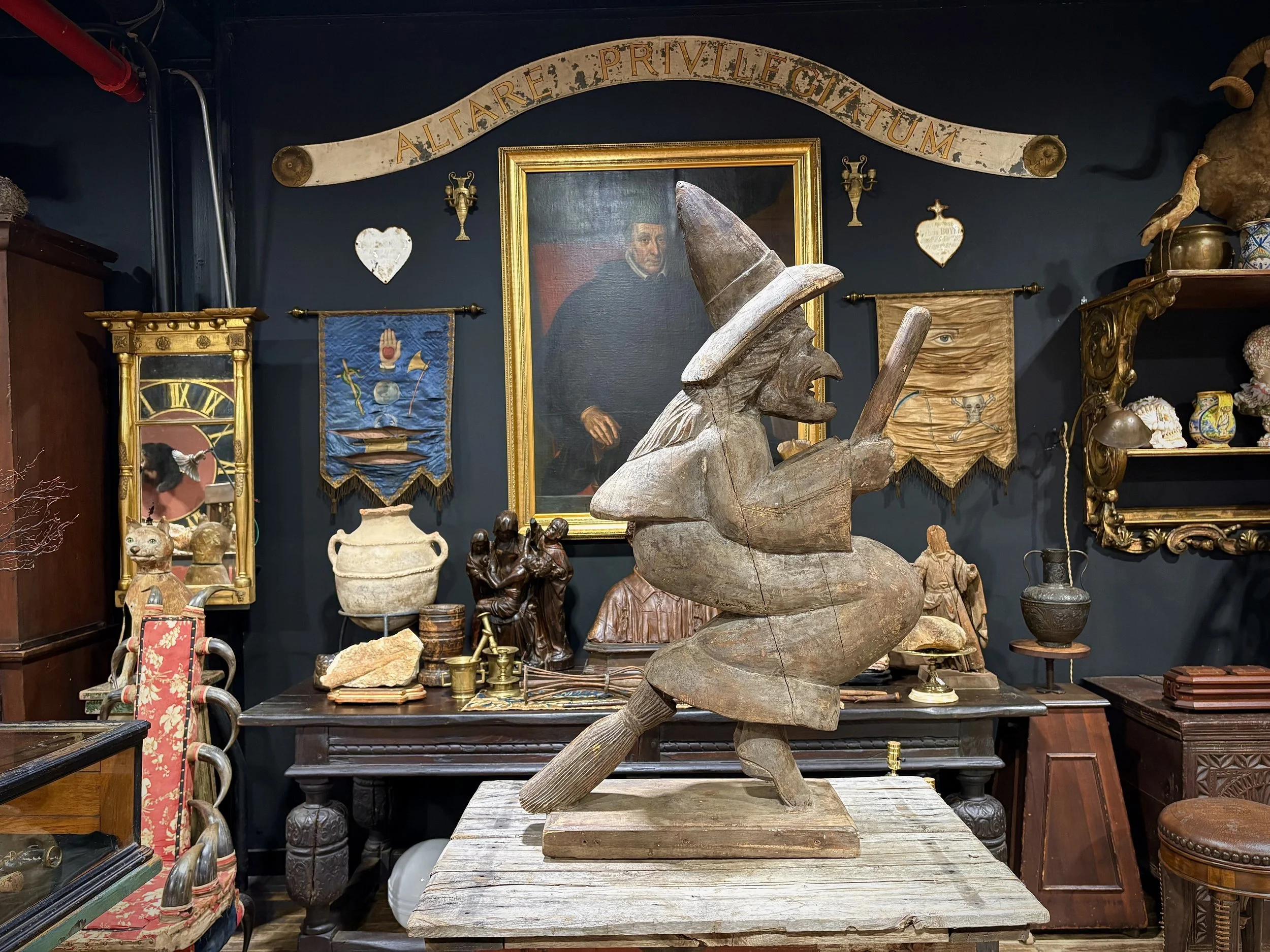 A wooden witch sculpture with a tall hat, holding a broom, displays in an antique shop surrounded by various vintage items and framed artwork on dark-colored walls.