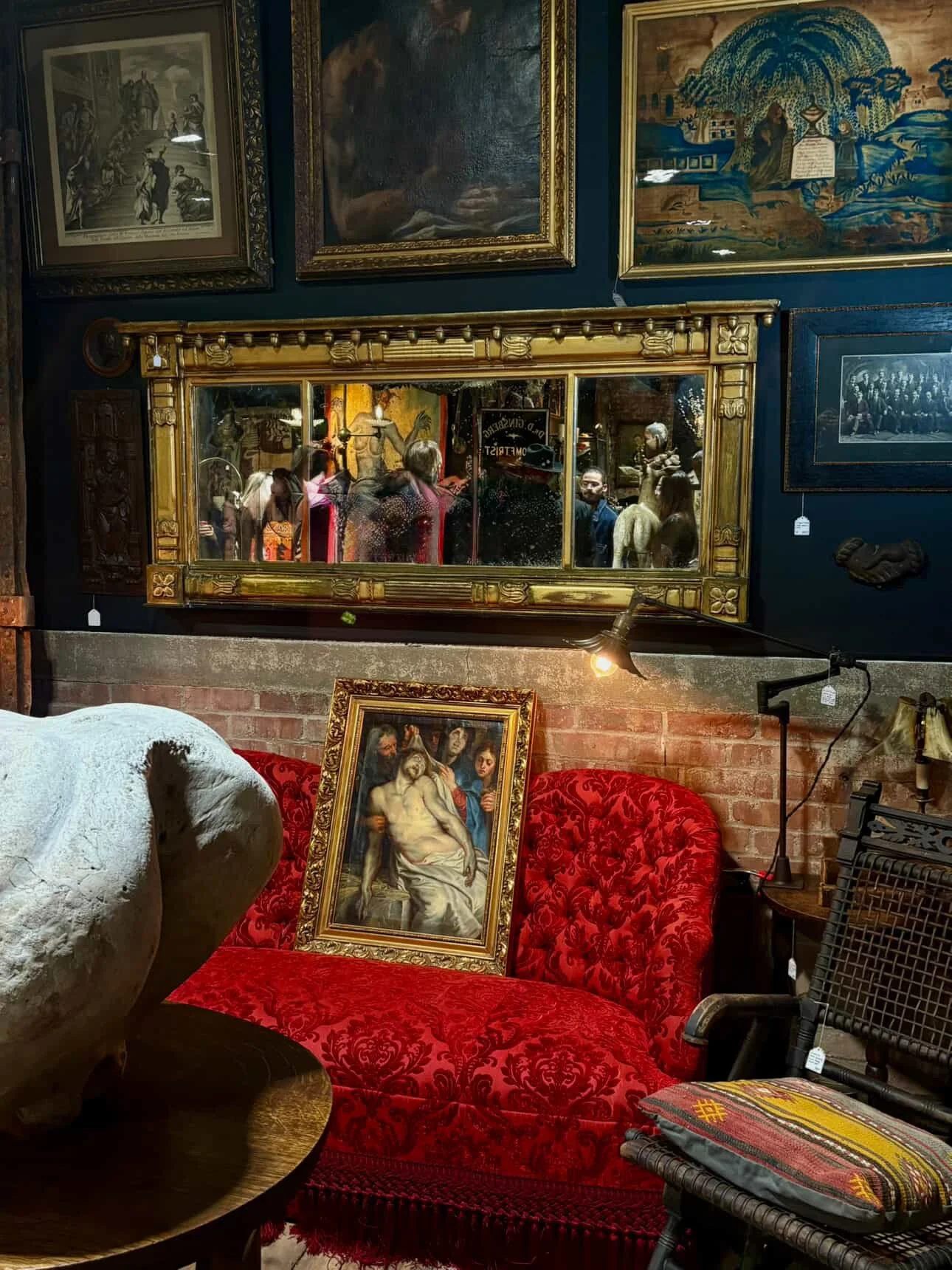 An art gallery or antique store interior with various paintings on dark walls, a large mirror, a red upholstered sofa with a framed painting of a classical scene resting on it, and a few visitors reflected in the mirror.