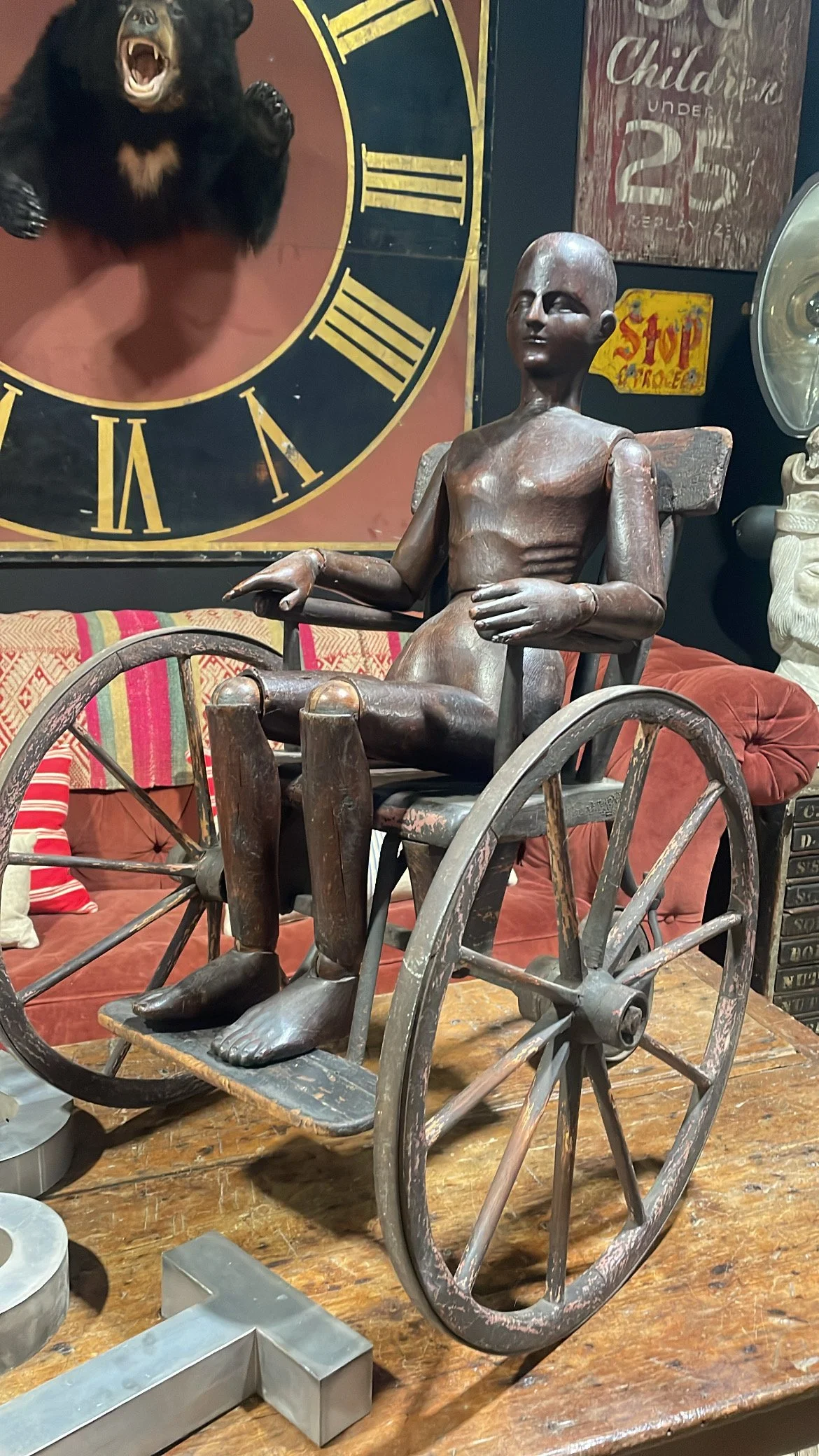 A vintage wooden statue of a seated human figure in a wheelchair with large wheels, positioned on a wooden table in a room decorated with various vintage signs and decor.