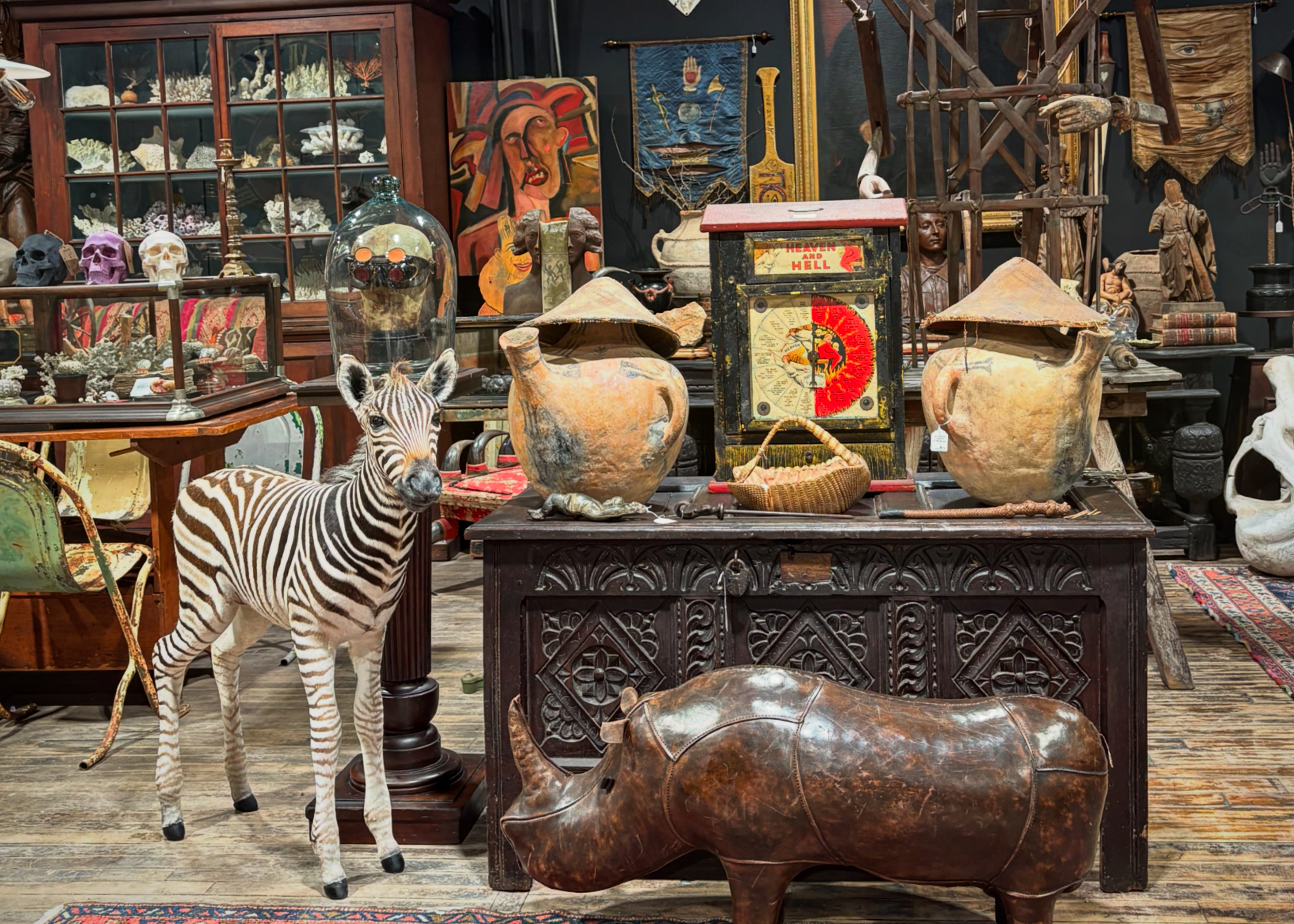 An antique store filled with vintage and rustic items, including a small zebra sculpture, large clay pots, a wooden display cabinet, sculptures, paintings, and decorative artifacts, all arranged on shelves and tables.